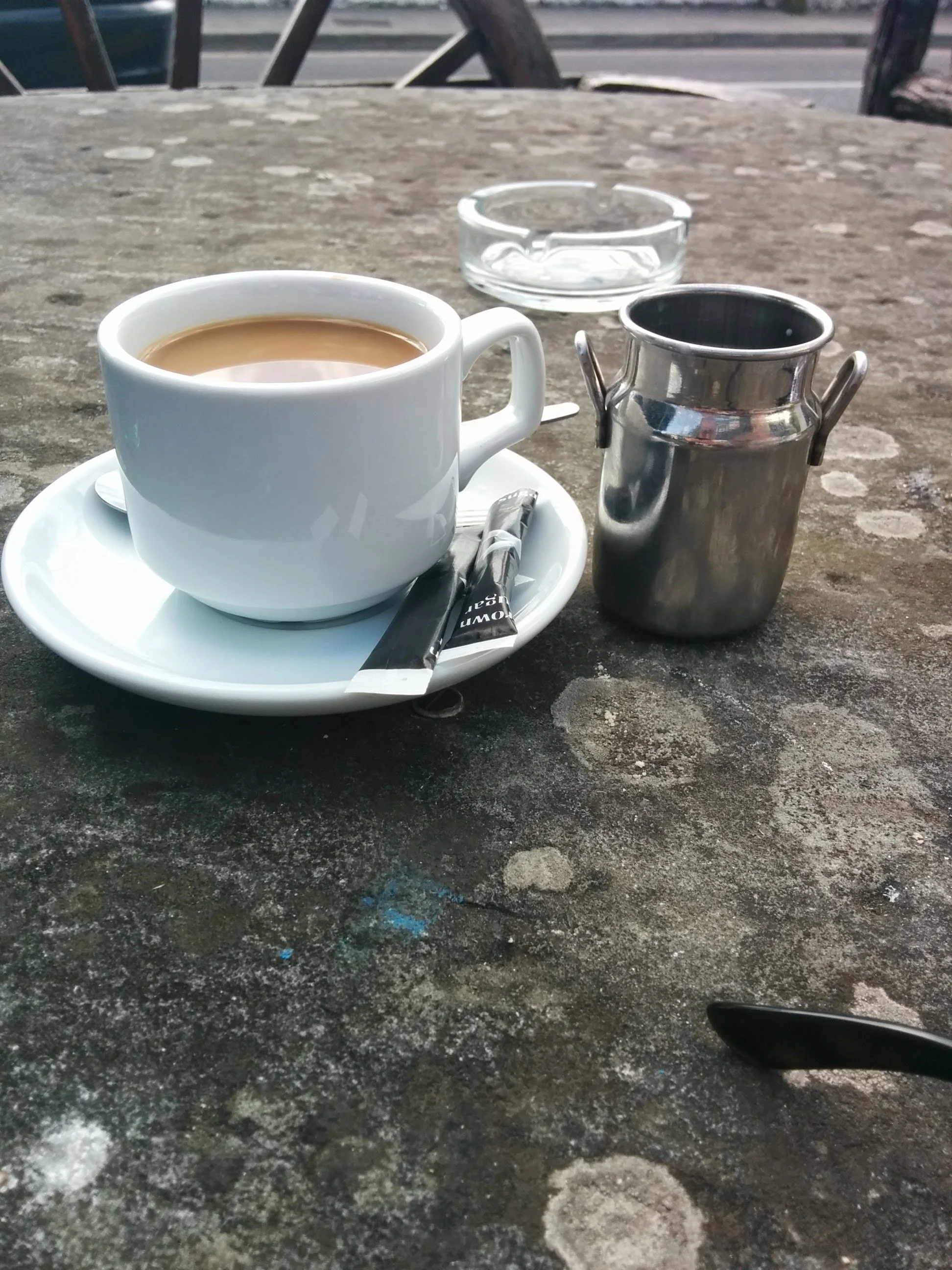 A white ceramic cup filled with coffee on a saucer with a wrapped spoon, a small metallic container, and an empty glass ashtray on a weathered dark surface table outdoors.