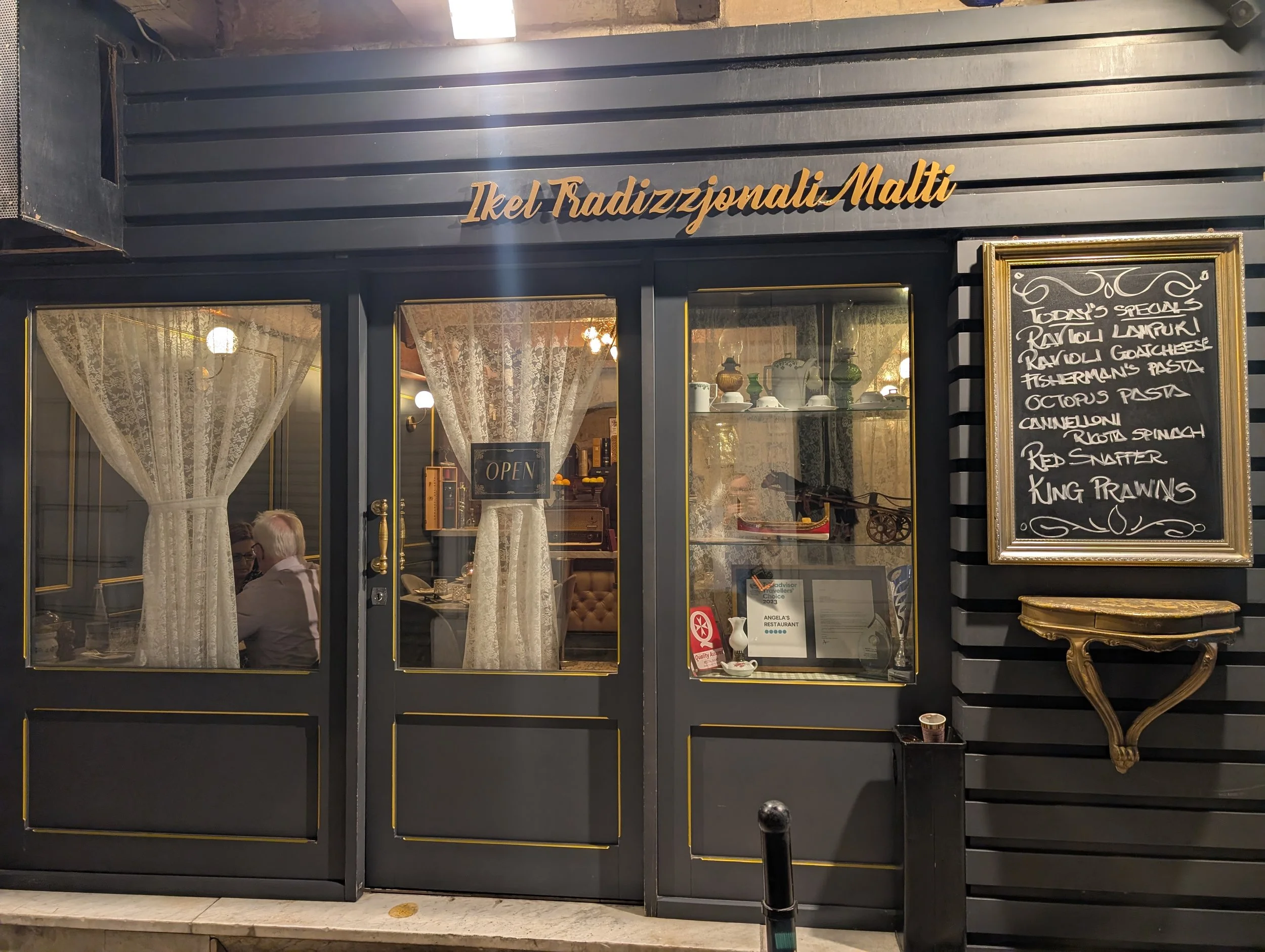 Exterior view of an Italian restaurant called 'Ikel Tradizzionali Malt.' The restaurant has a black facade with gold accents, lace curtains in the windows, and a chalkboard menu on the side listing various Italian dishes.
