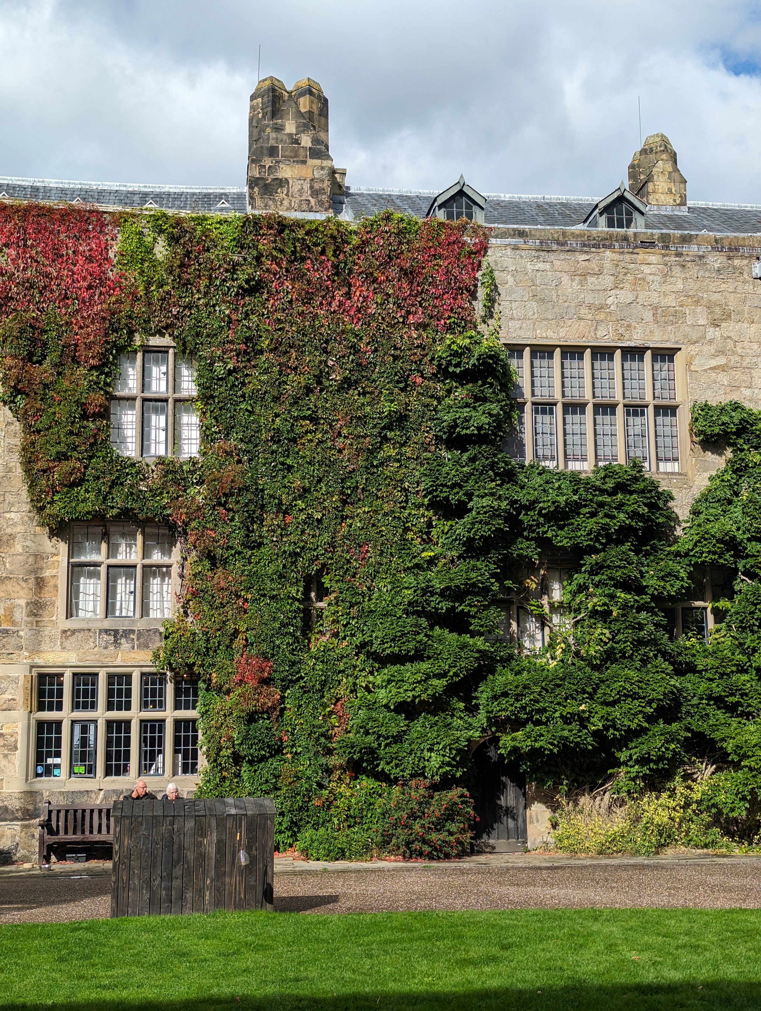Stone building covered with leafy green and red ivy, featuring large multi-pane windows, with a small park and two people sitting on a bench in front, and a trash bin nearby.