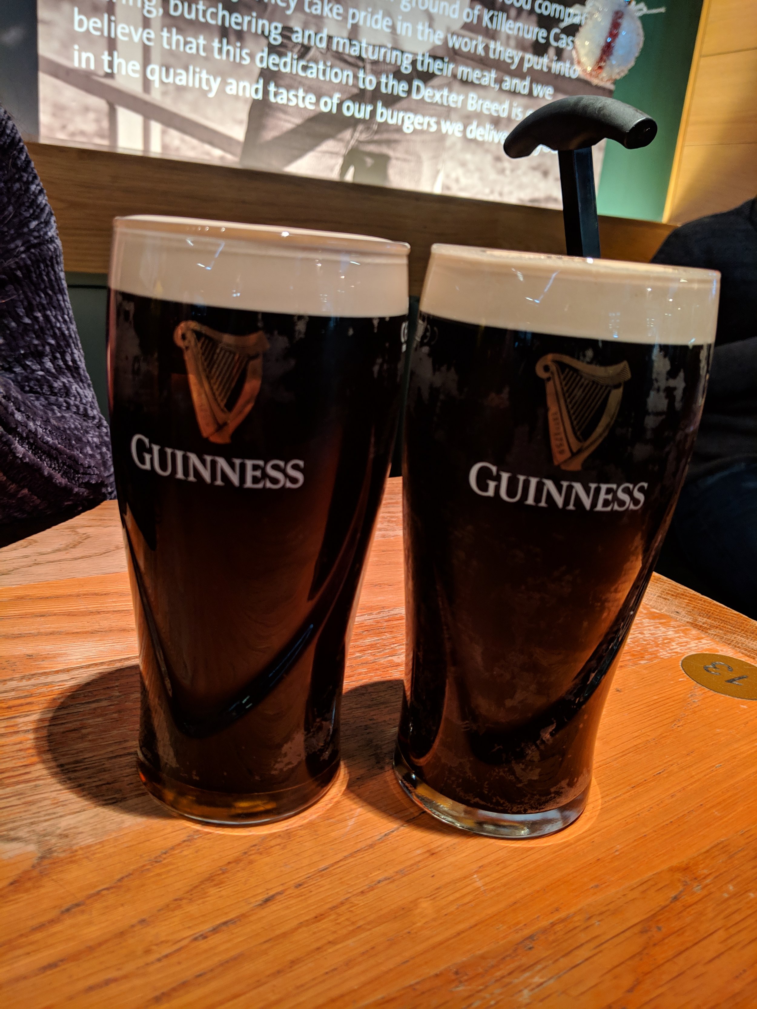 Two Guinness beer glasses filled with dark stout beer and topped with foam, placed on a wooden table in a pub or restaurant.