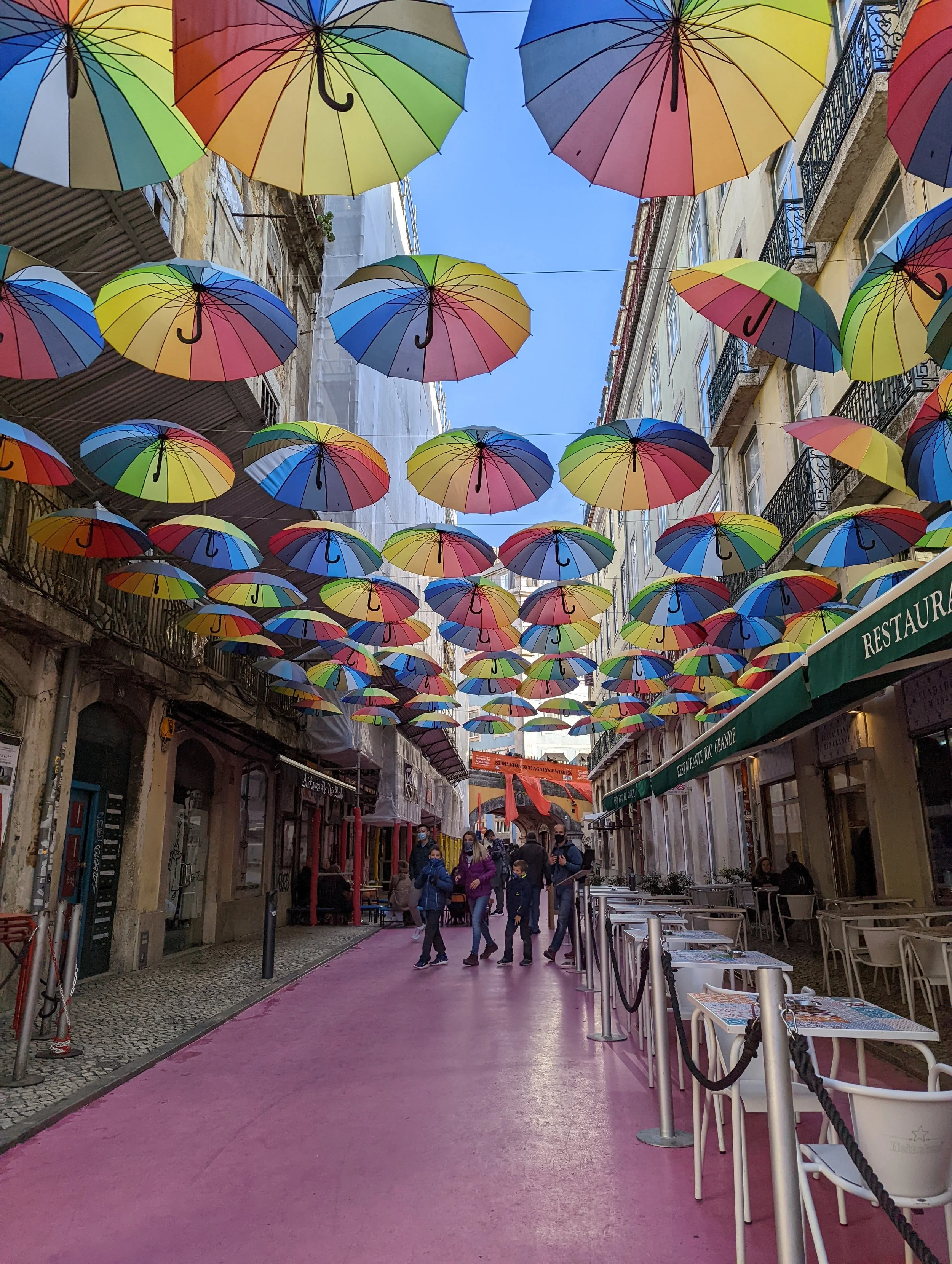 Colorful umbrellas hanging overhead in a street, creating a vibrant canopy with people walking below and outdoor tables on the side.