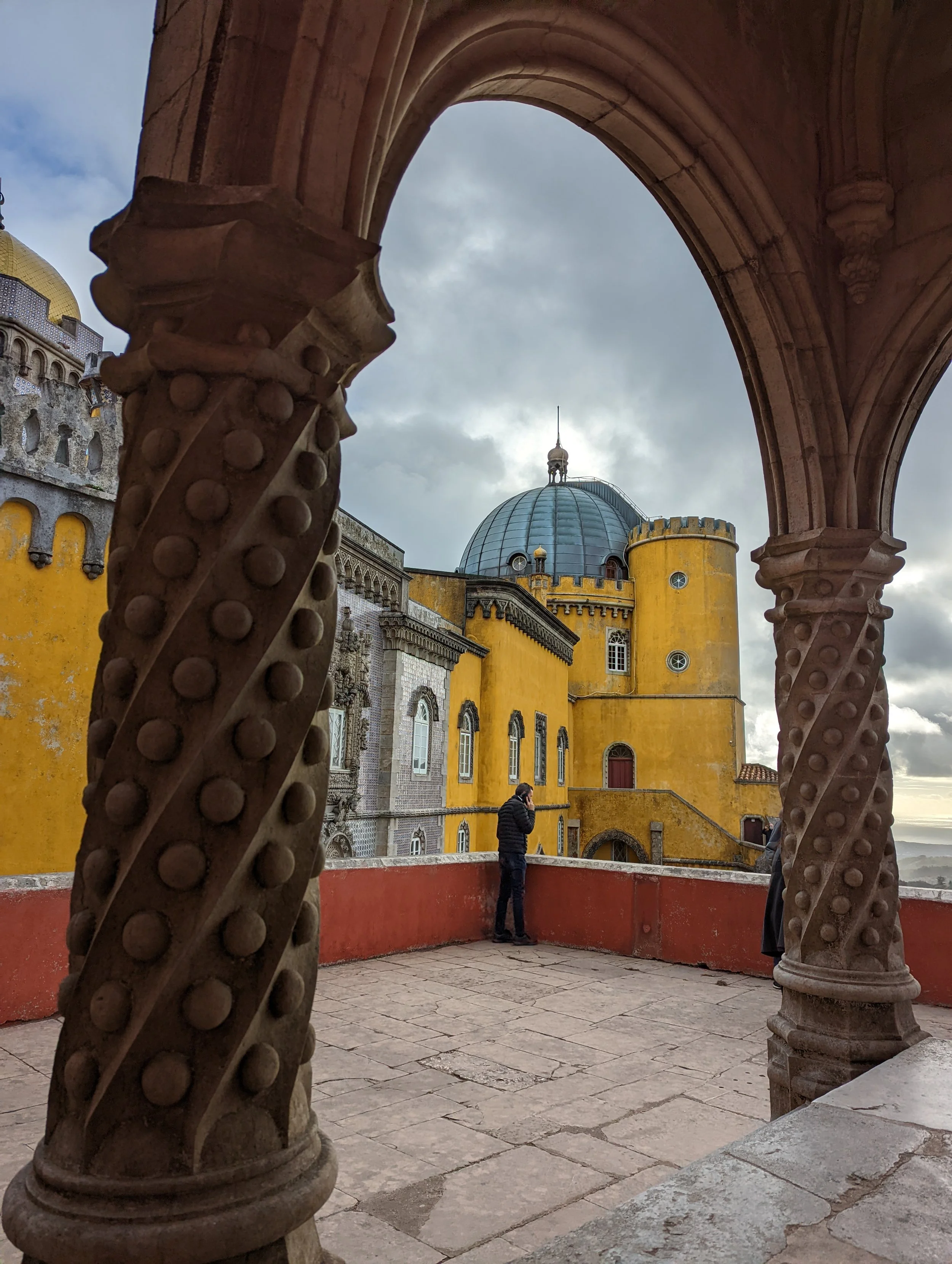 View through stone arches of a yellow castle with a blue domed roof, small windows, and a tower with a flag, against cloudy sky.