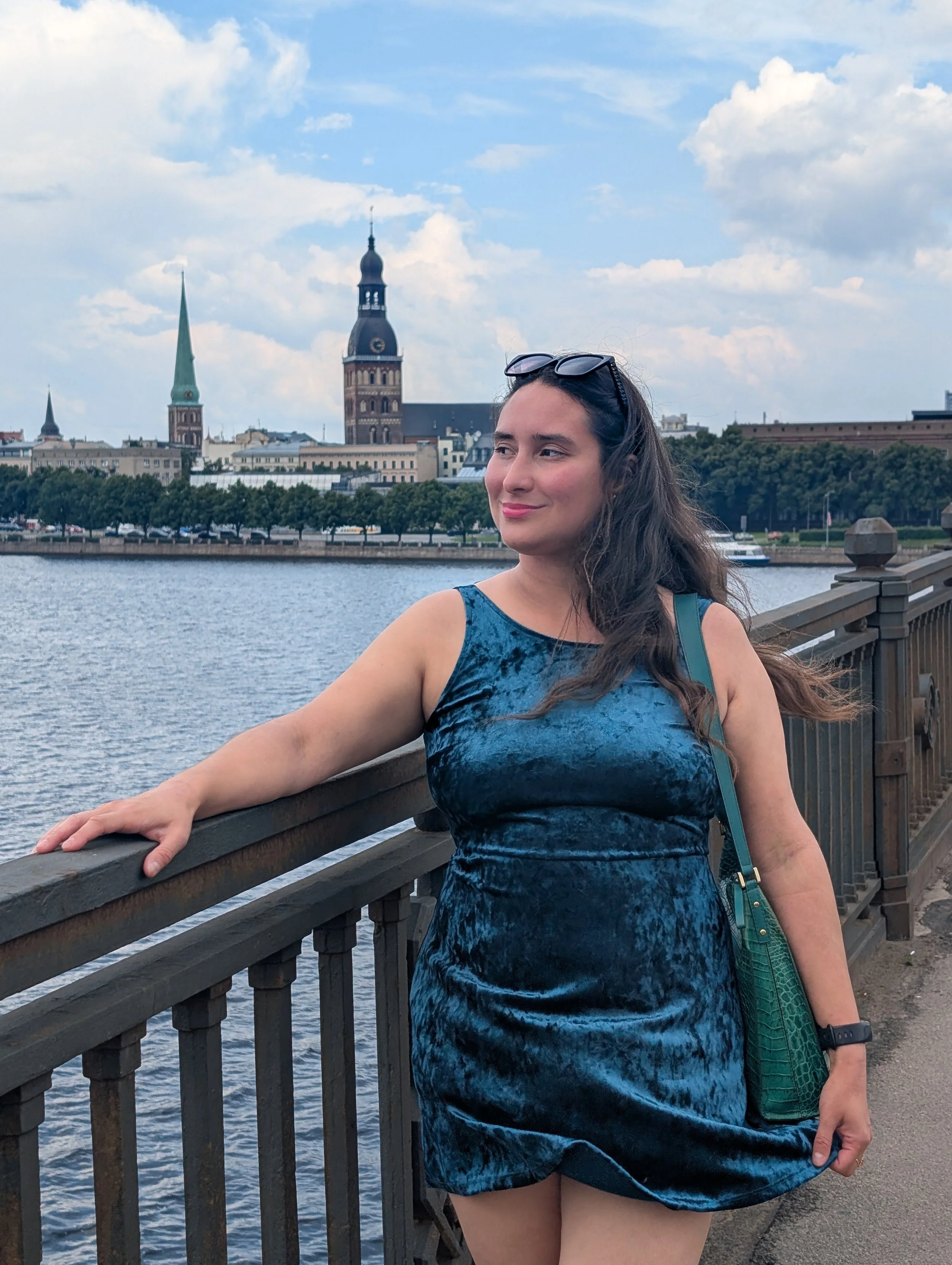 A young woman in a blue dress standing on a bridge over a river with historic church towers and a cityscape in the background, wearing sunglasses on her head and a green purse.