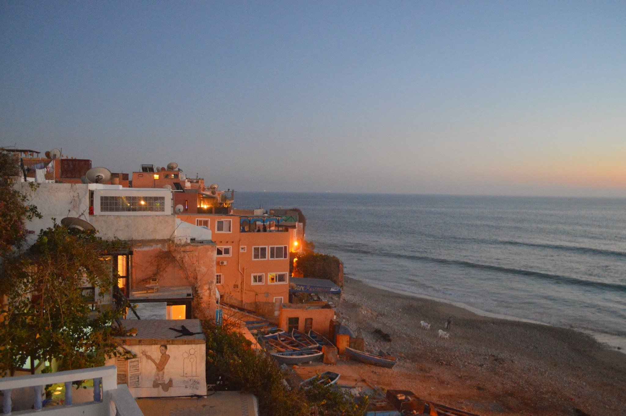 Sunset over a coastal town with colourful buildings, satellite dishes on rooftops, and boats on a pebbled beach.
