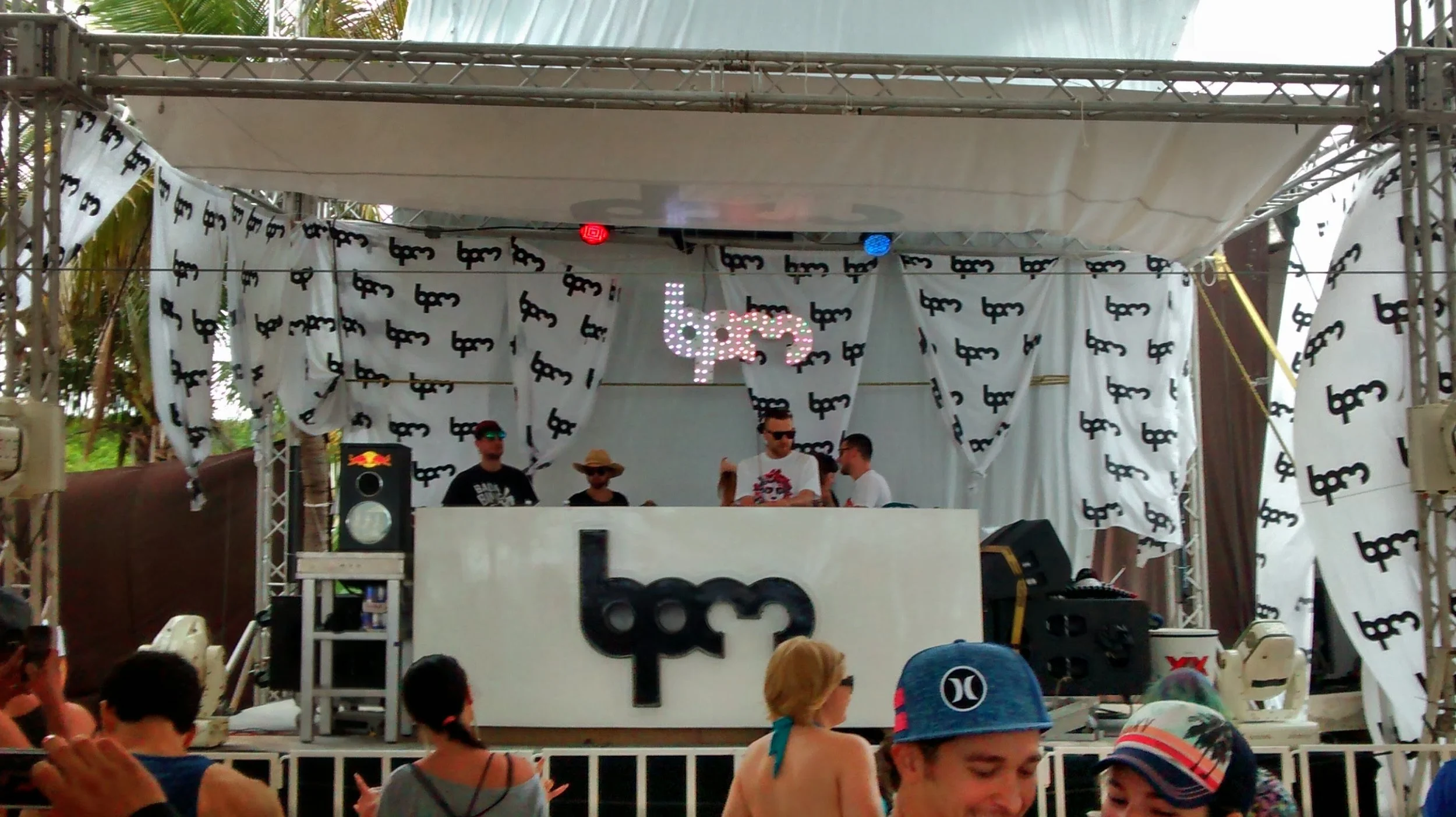 A DJ performing on a stage decorated with black dollar signs on white fabric, with a crowd watching in front. There are some people on the stage, one wearing sunglasses and another with a sun hat, and a colorful LED sign displaying 'bpm' in the backg
