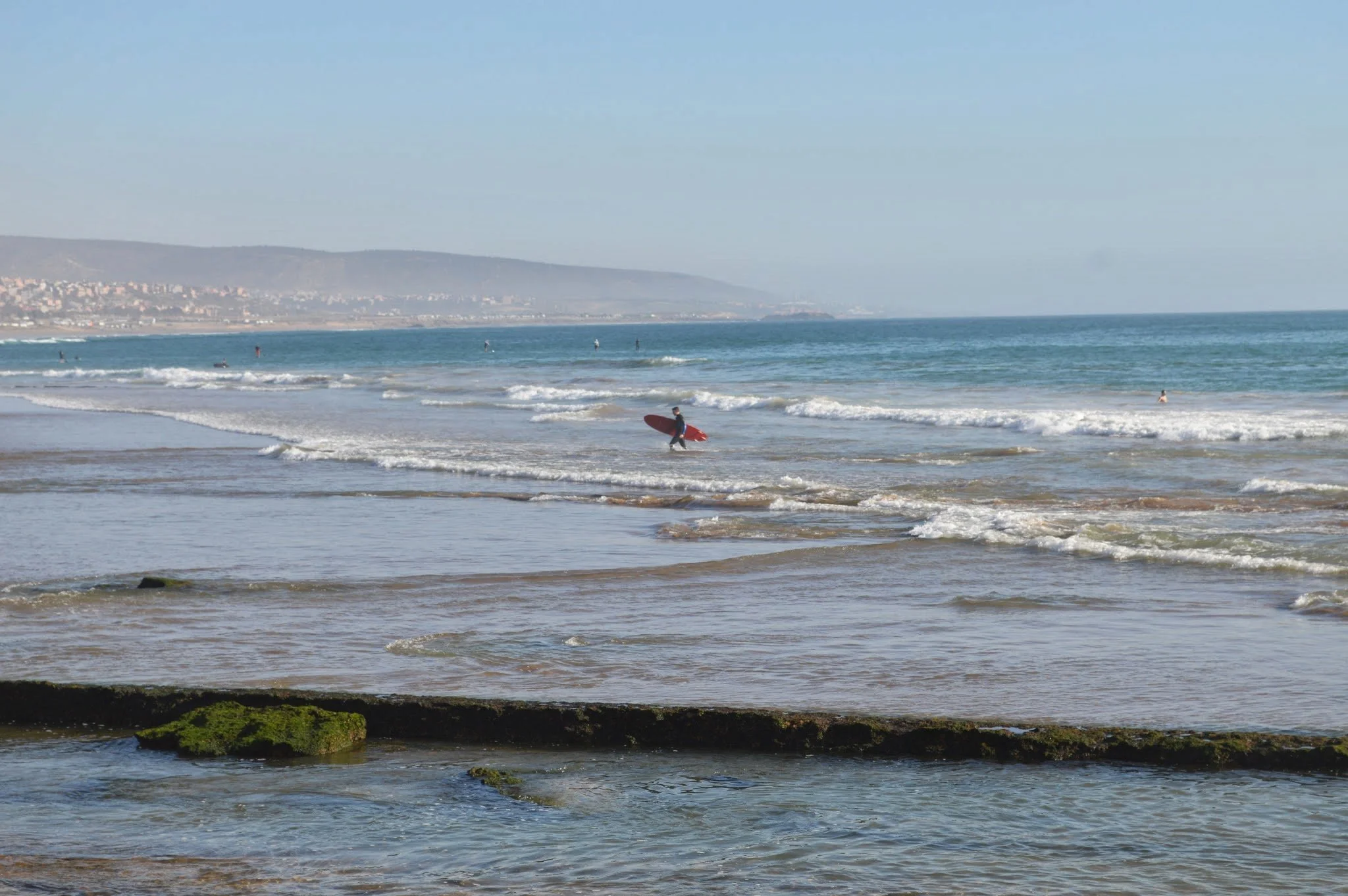 A beach scene with a person carrying a surfboard walking near the water, small waves, rocks with green moss in the foreground, and a distant shoreline with hills.
