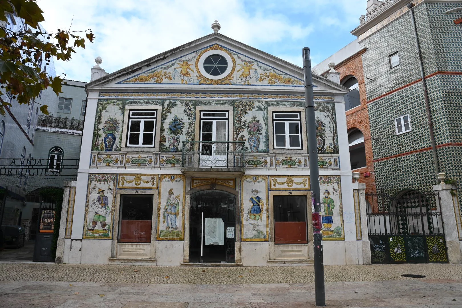 Frescoed building with decorative murals and floral paintings, features three windows on the upper floor, entrance on the ground floor, and a small balcony in the middle. Surrounding buildings are modern, with one having a tiled patterned facade.