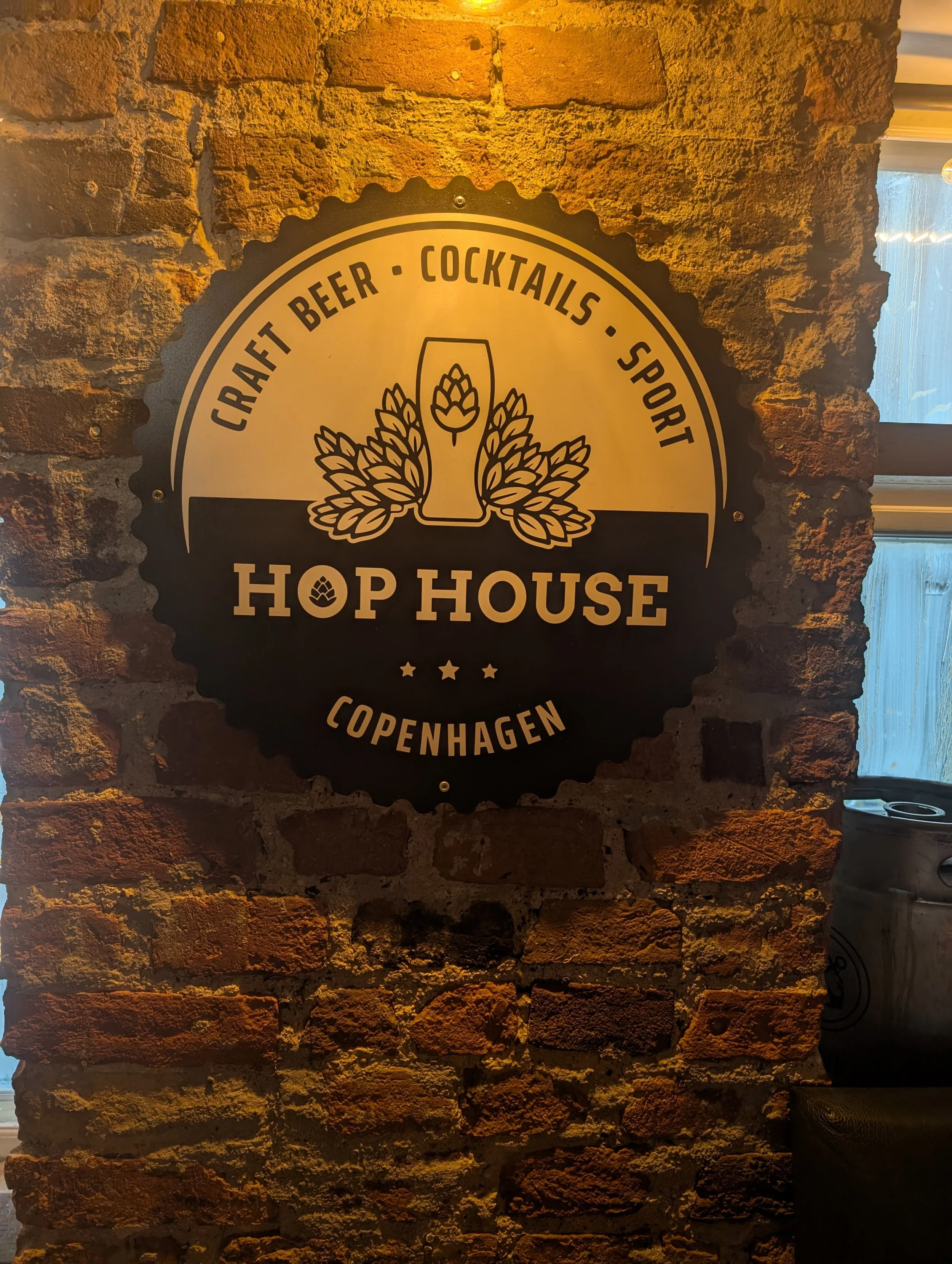 Sign on brick wall featuring a beer glass with hop and barley decorations, advertising craft beer, cocktails, sport, and Copenhagen at Hop House.