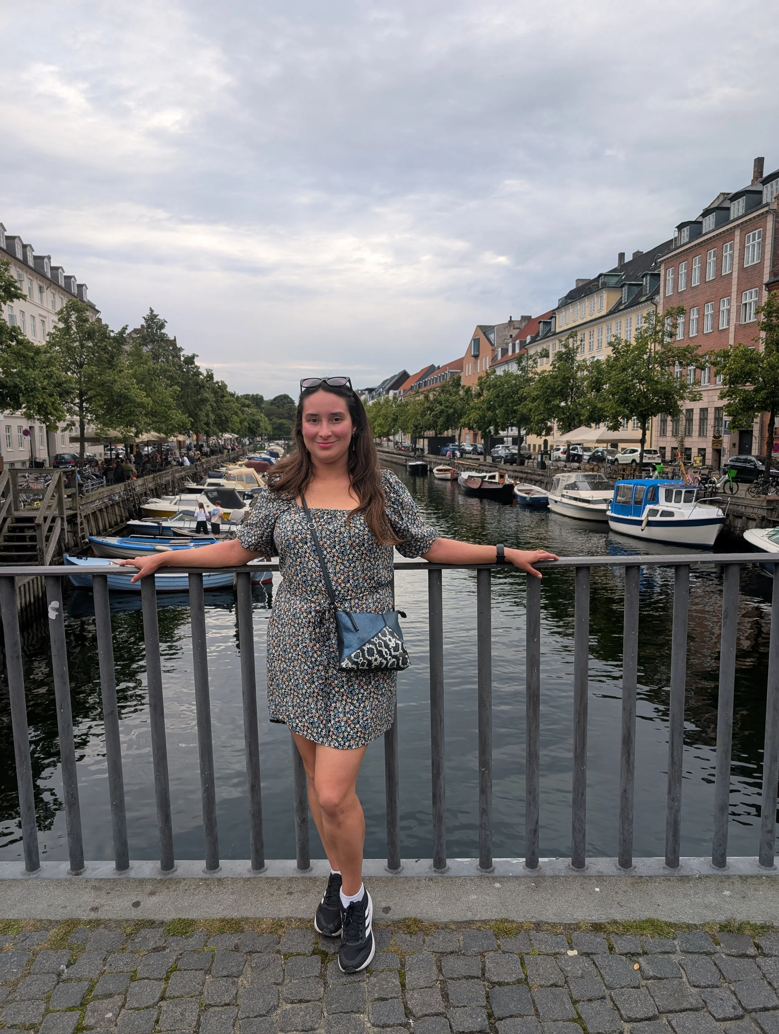 A young woman with long brown hair, wearing a floral dress, black sneakers, and sunglasses on her head, stands on a bridge with her arms resting on the railing, in front of a canal lined with boats and colorful buildings under a cloudy sky.
