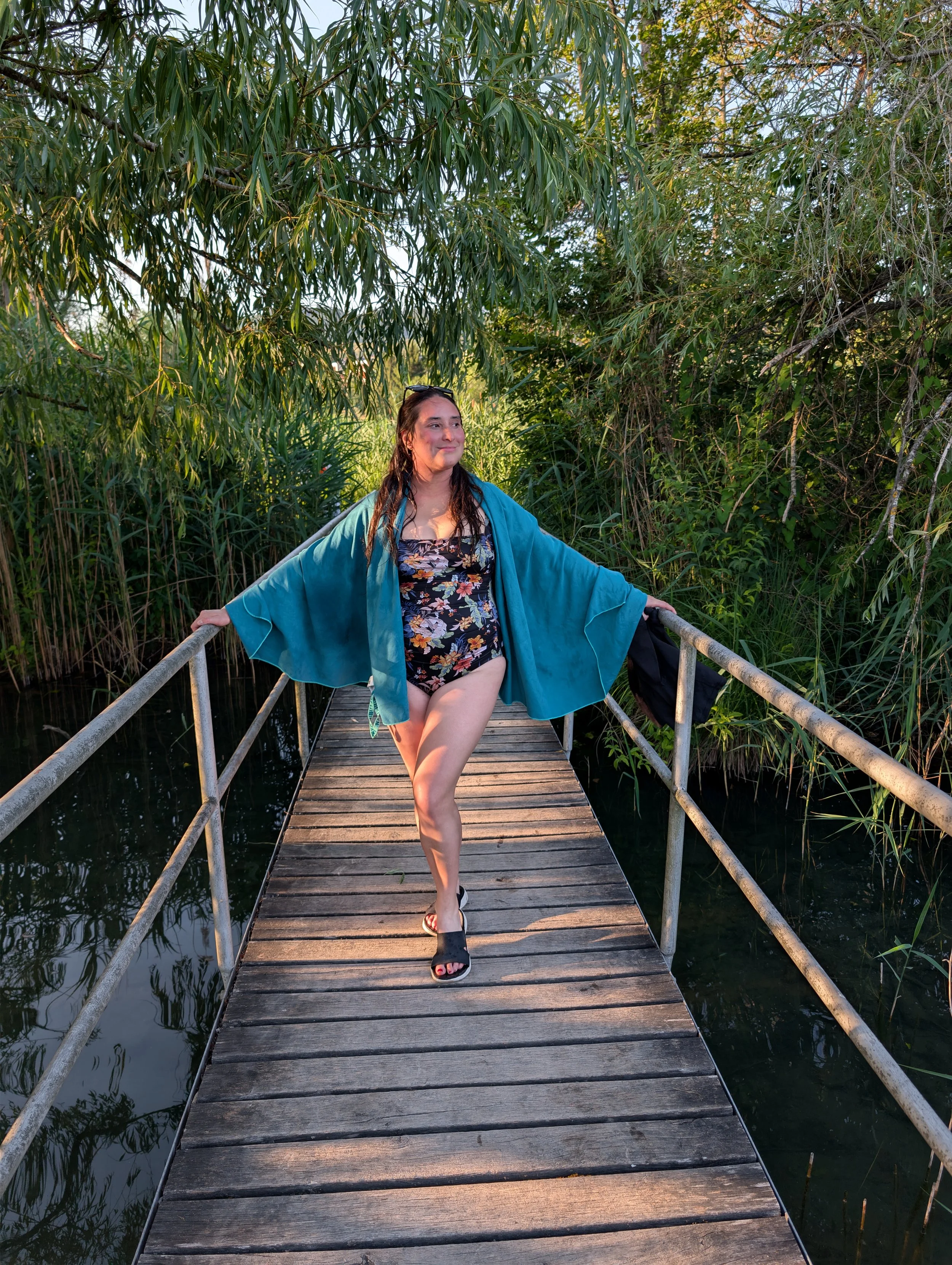 A woman in a floral swimsuit and blue cover-up standing on a wooden boardwalk surrounded by lush greenery and water, with her arms outstretched and a smile on her face.