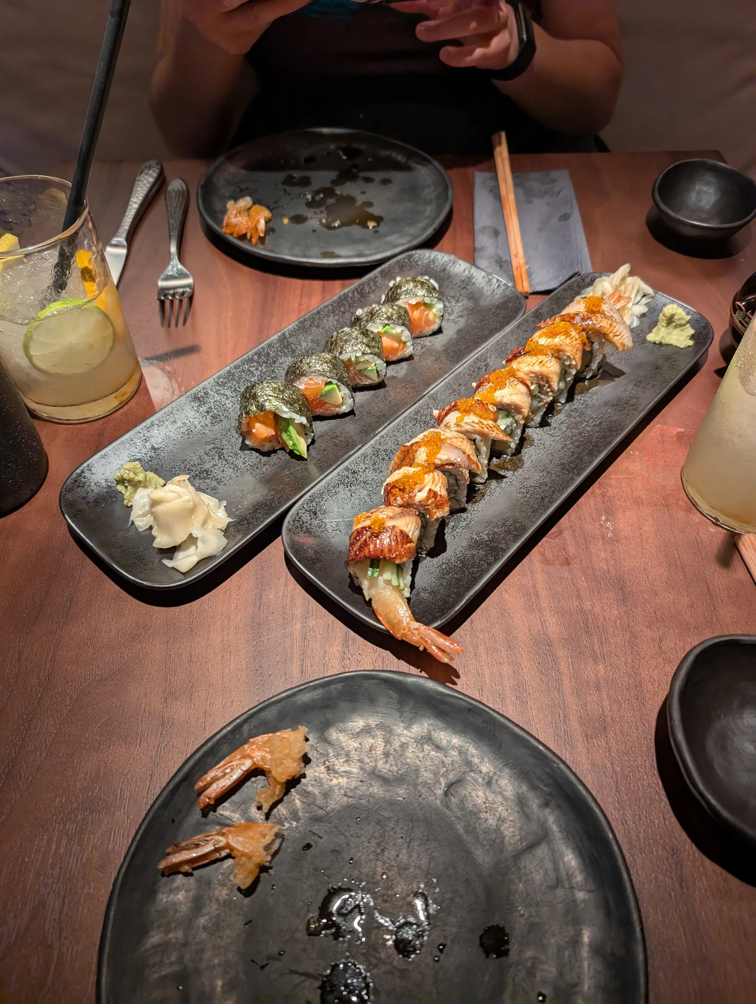 A dining table with two plates of sushi, one with salmon, avocado, and seaweed, and the other with cooked fish and fish roe; a drink with lemon and lime slices, a black bowl, and utensils; someone taking a picture of the sushi with their phone.