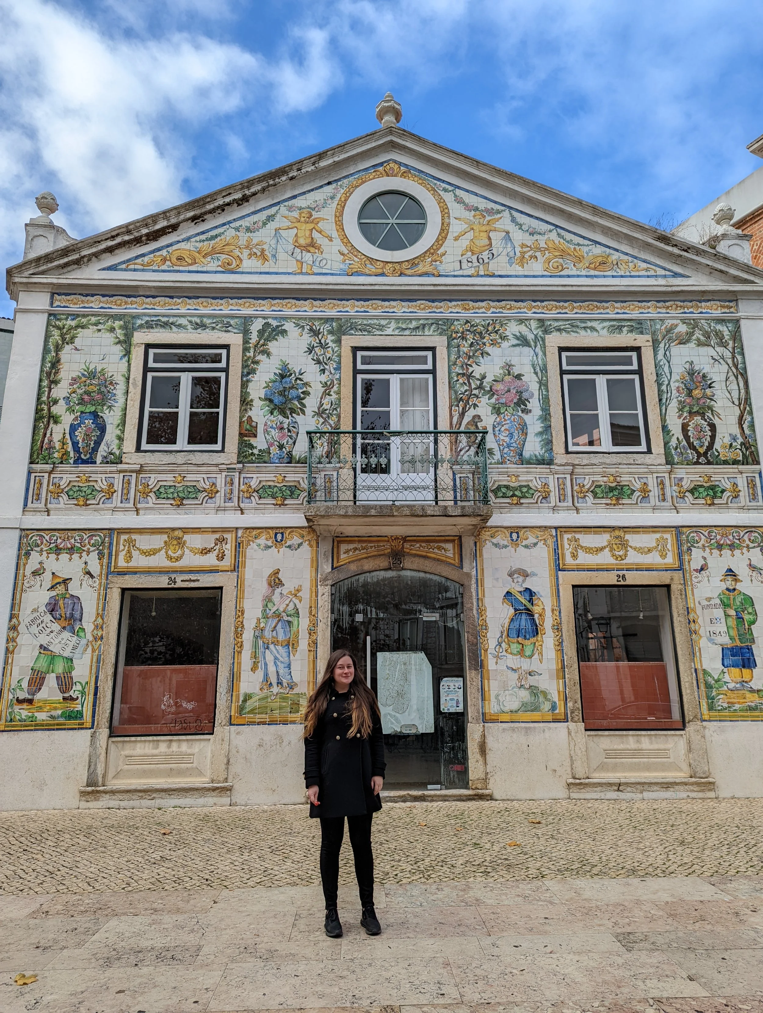 A young woman standing in front of a building with colorful ceramic tile art depicting people, flowers, and decorative elements. The building's facade features windows, a small balcony, and a decorative triangular pediment with the year 1865 and two 