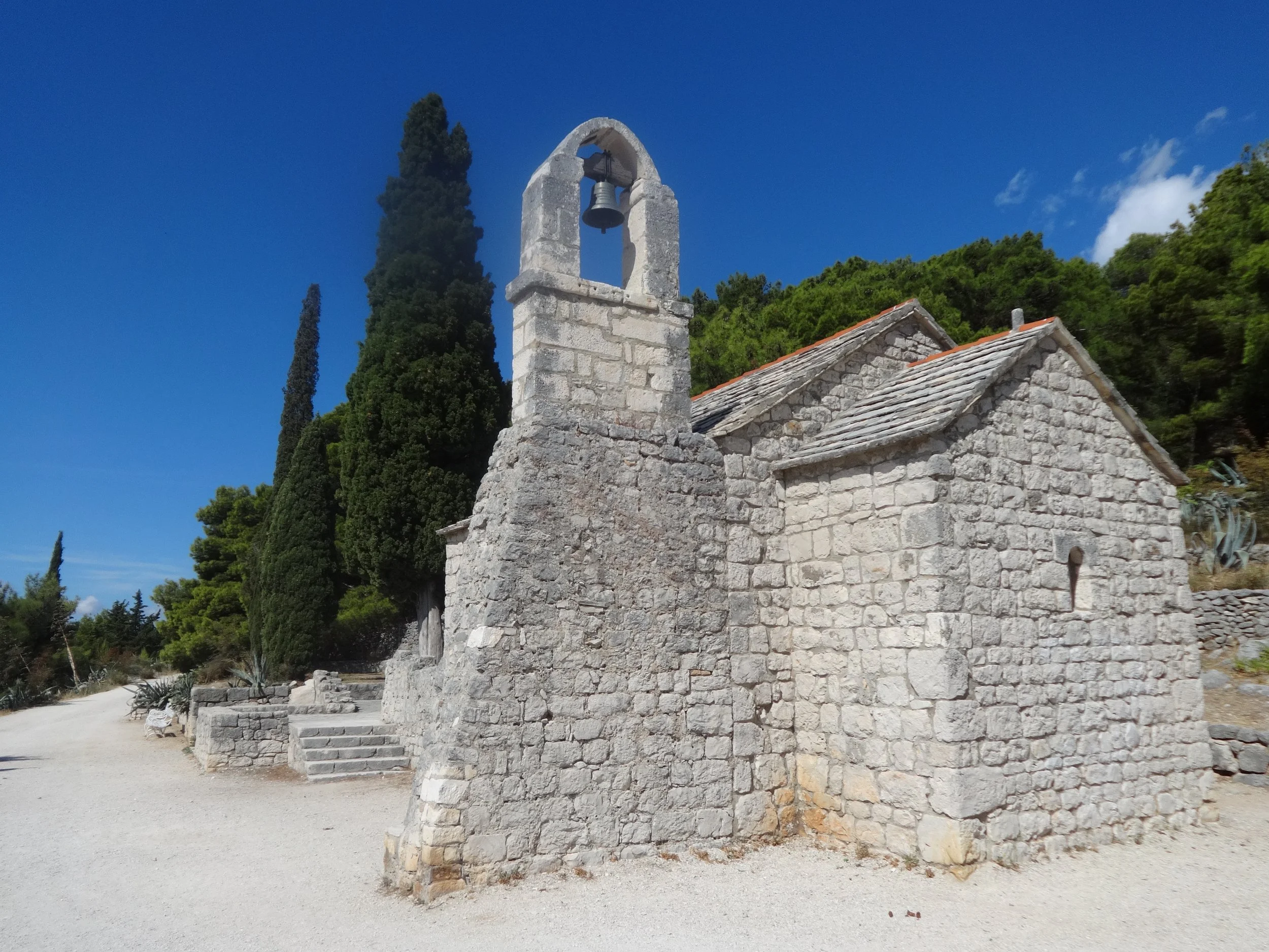 Stone building with a bell tower and red-tiled roof, surrounded by green trees and a clear blue sky.