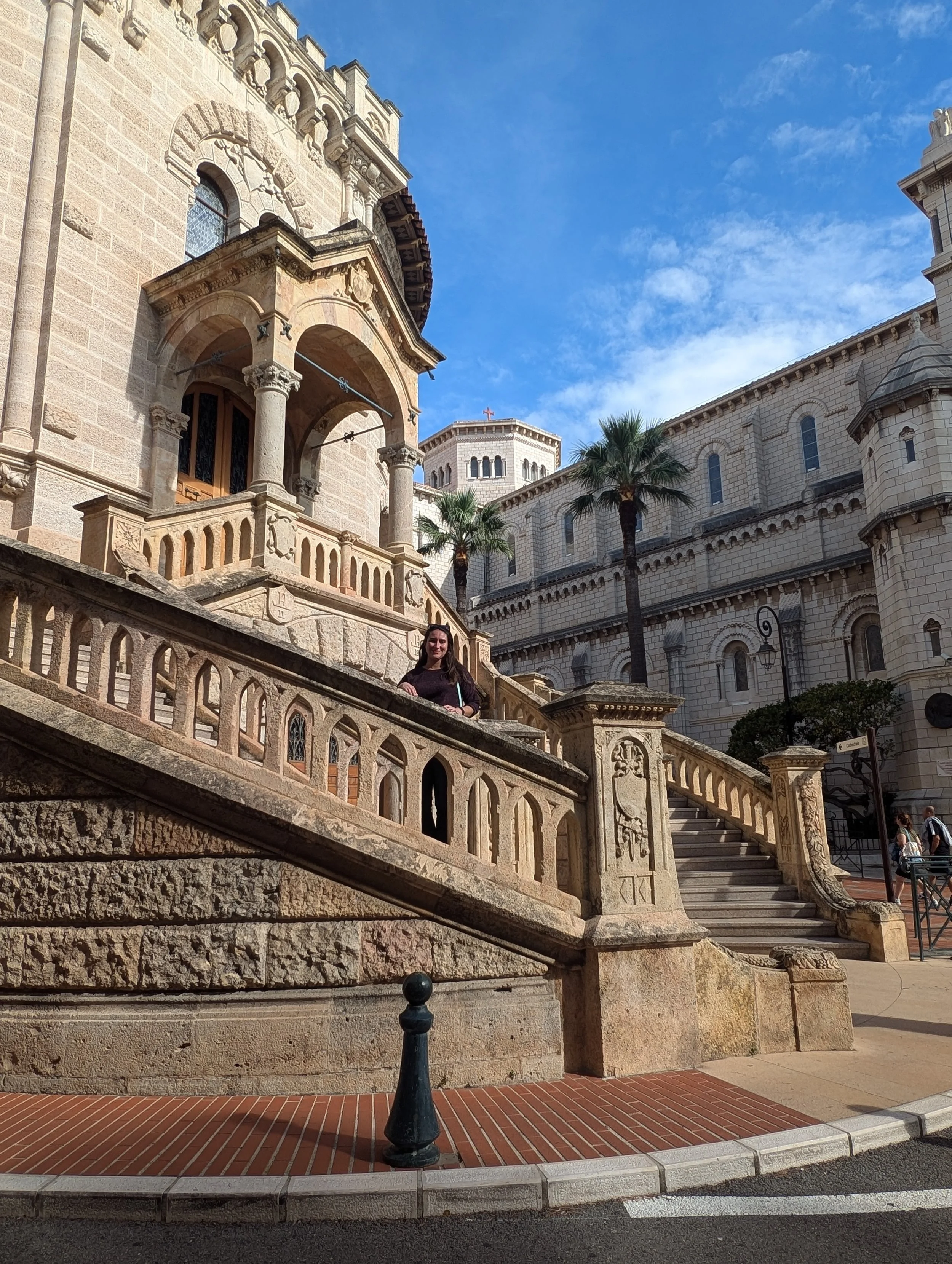 A woman standing on a stone staircase in front of a historic stone building with arched windows, columns, and a balcony with decorative railing, under a partly cloudy blue sky with palm trees nearby.