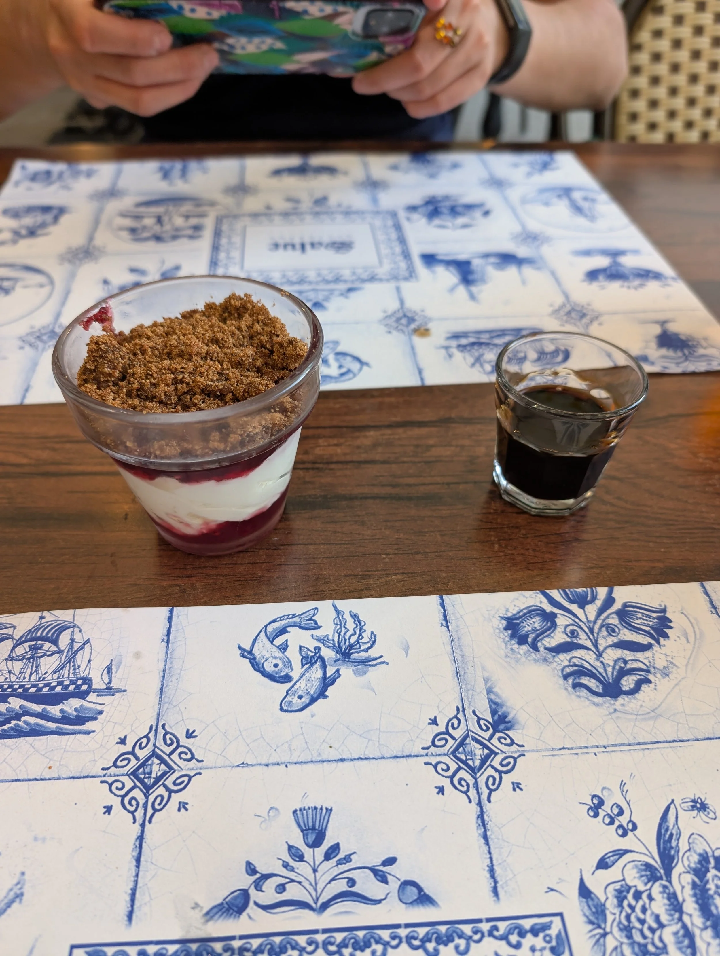 A table with a blue and white tile pattern tablecloth, featuring various artistic designs including animals and decorative elements. In the foreground, there is a dessert in a glass with layers of cream and berry sauce topped with brown crumbs, and a