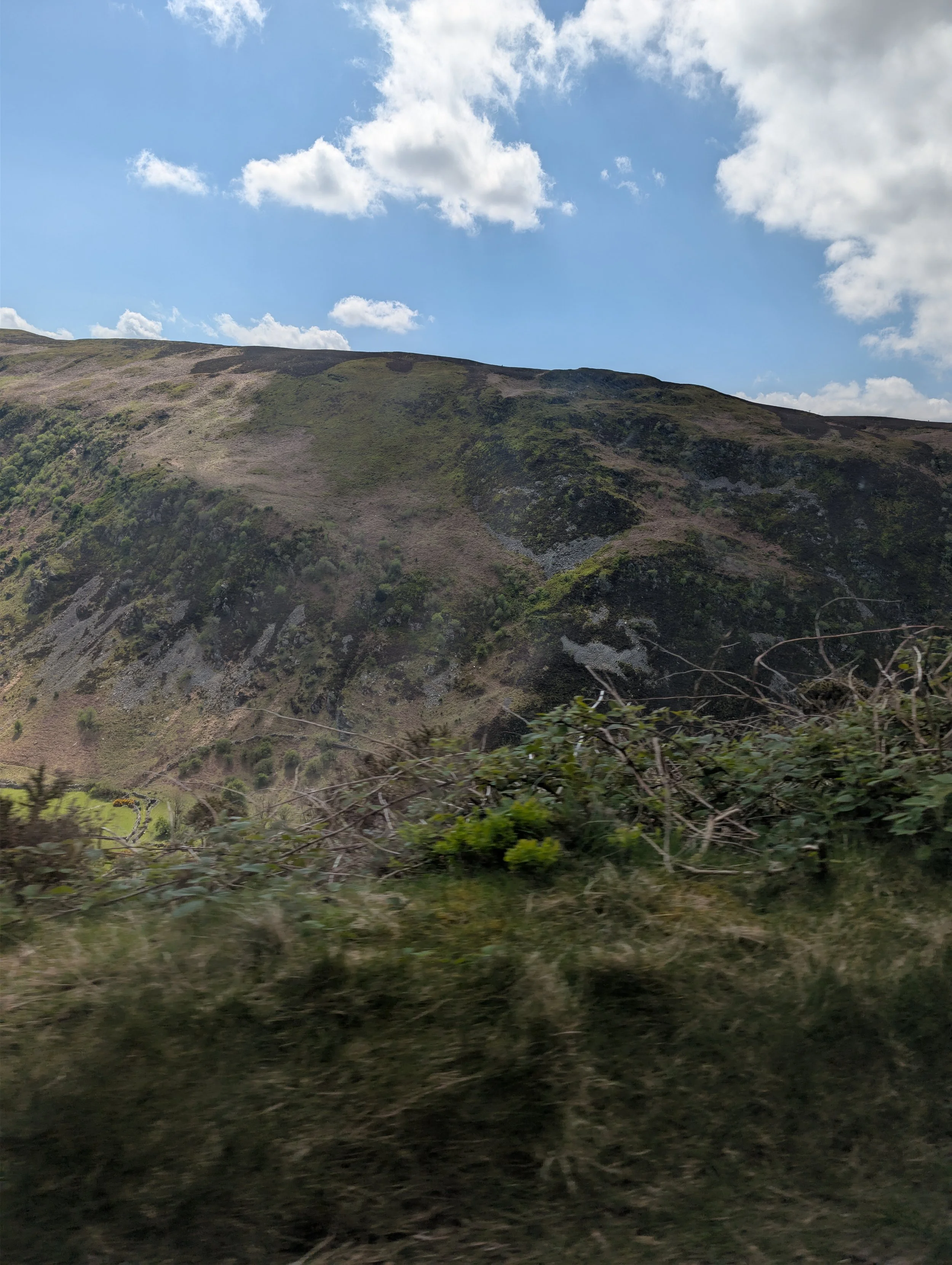 A rugged hillside with patches of green and rocky terrain under a bright blue sky with fluffy white clouds.