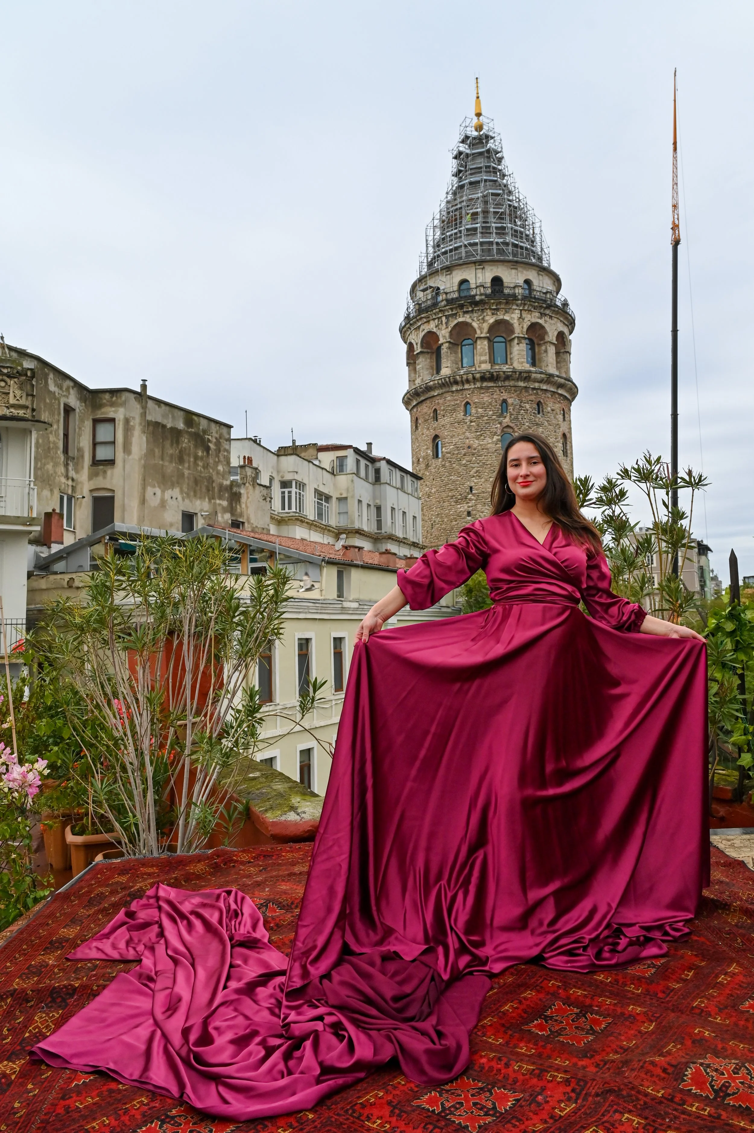 A woman in a long, elegant, satin burgundy dress posed on a red patterned Persian rug on a terrace. Behind her, the Galata Tower with scaffolding, and other historic buildings are visible under a cloudy sky. She is smiling confidently, holding out th