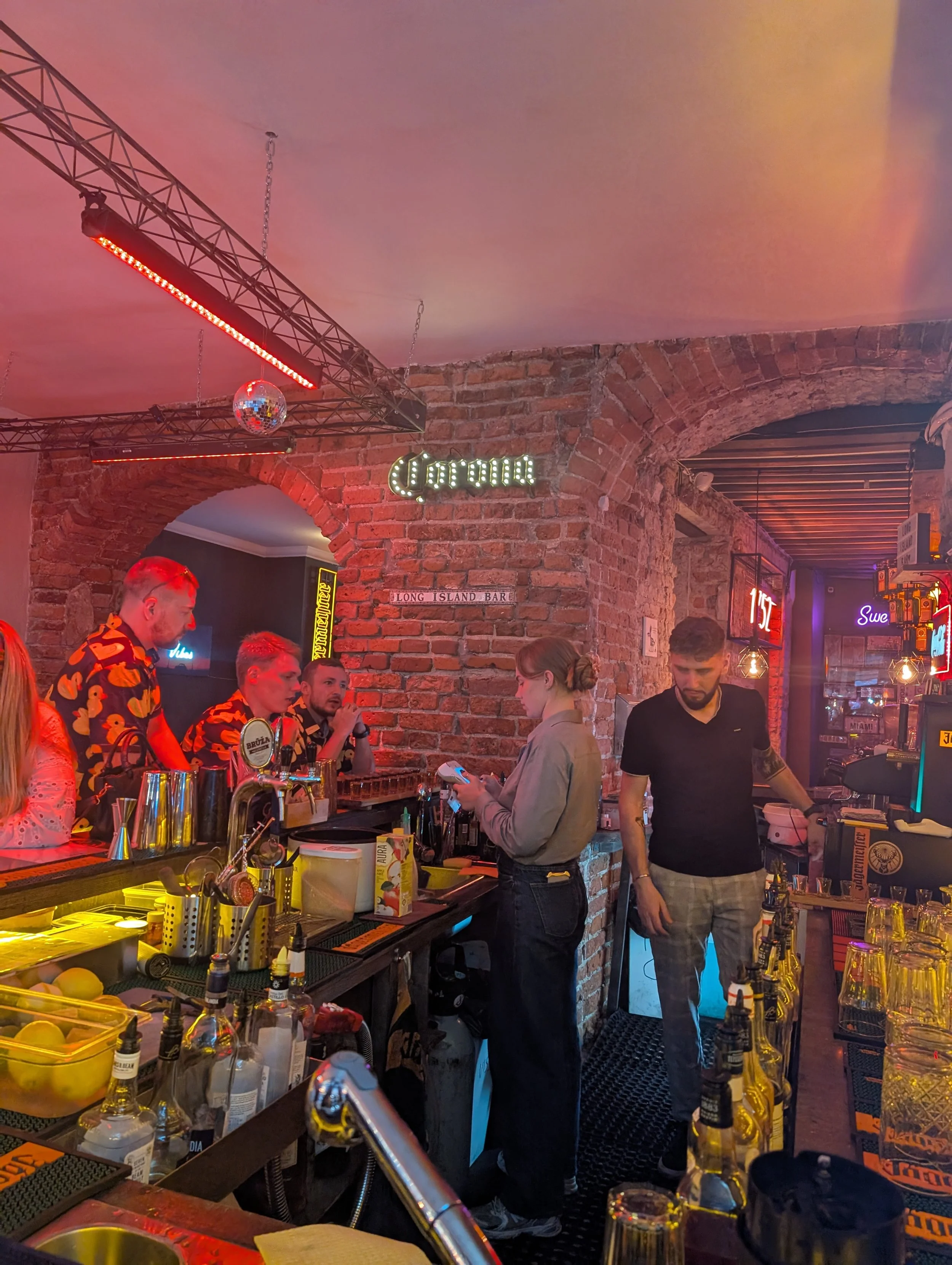 People gathered at a bar with exposed brick walls, neon signs, and a disco ball, engaged in conversation and preparing drinks in a lively nightlife setting.