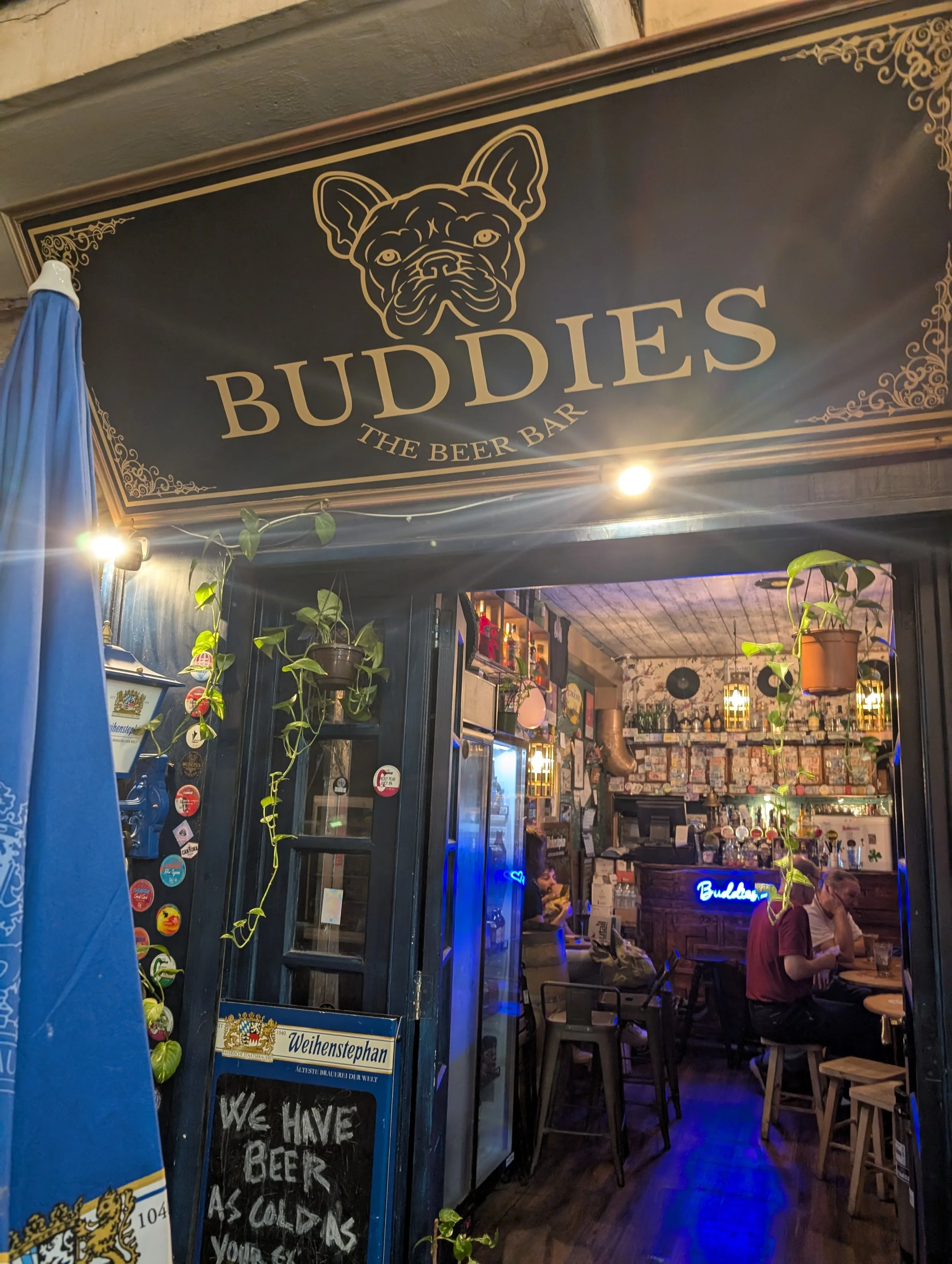 Entrance to a bar named Buddy's The Beer Bar featuring a sign with a French Bulldog illustration. Inside, there are decorative plants, a chalkboard with a beer-related message, and patrons seated at tables.