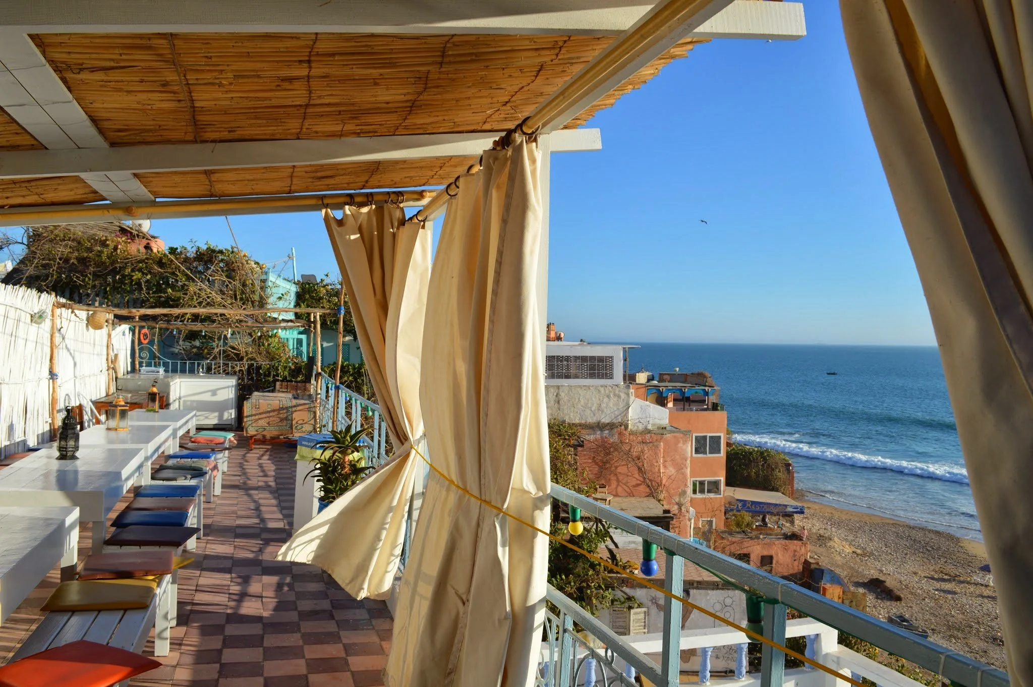 A balcony overlooking the ocean with white tables and colourful chairs, beige curtains, a bamboo roof cover, and nearby buildings with the ocean in the background under a clear blue sky.