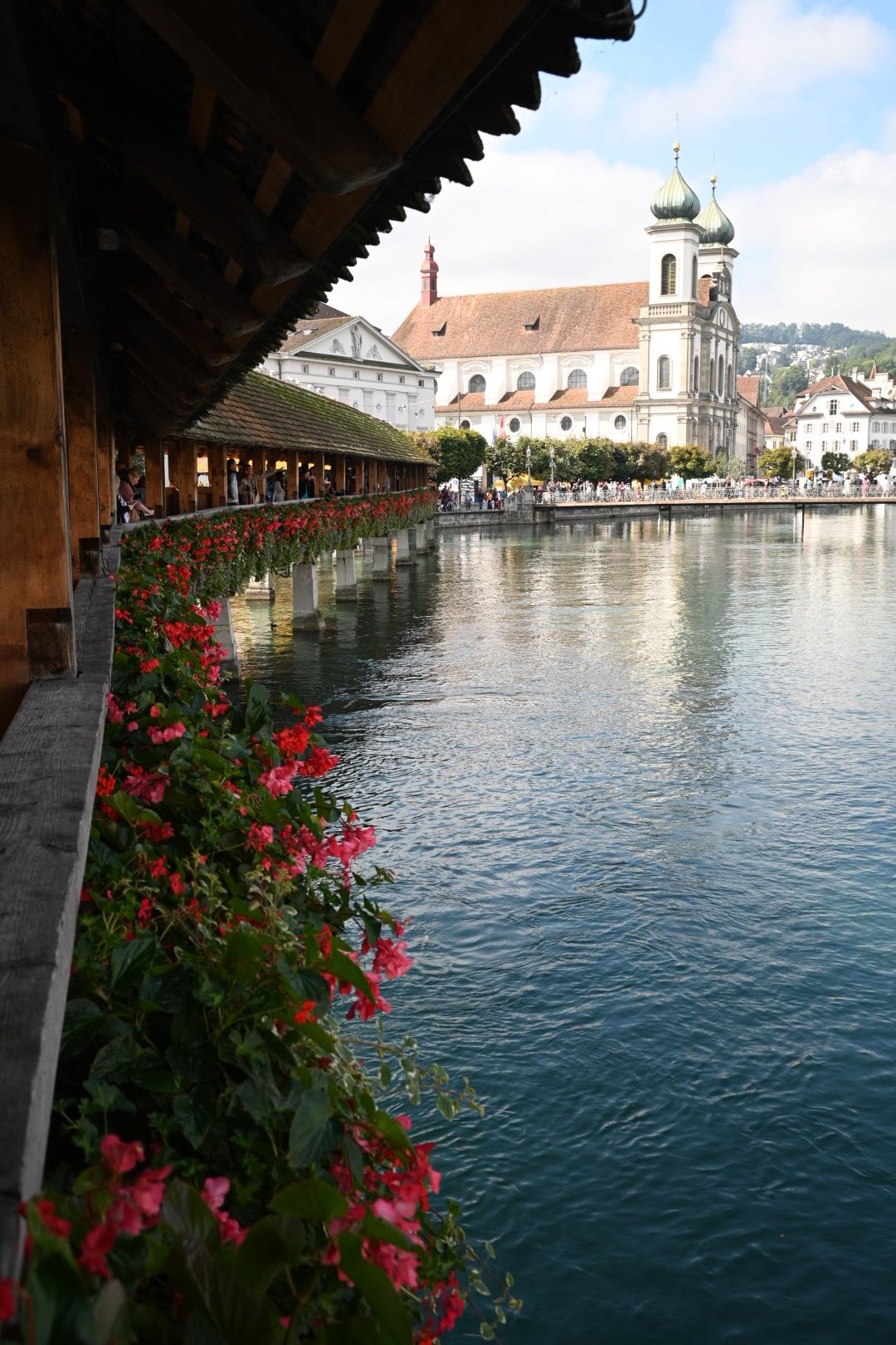 View of historic city with church and a bridge over a lake, flowers along the bridge, and people walking.