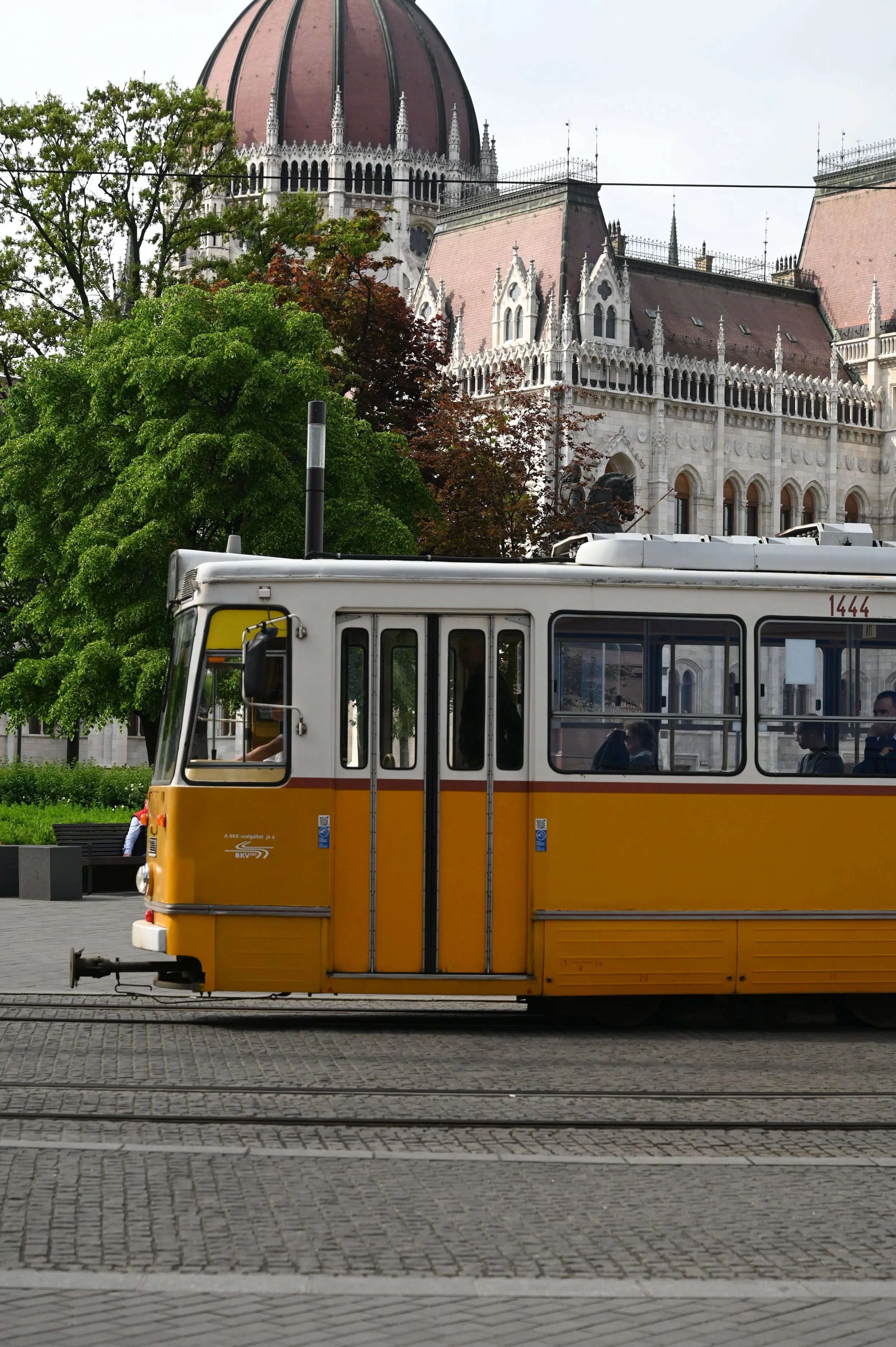 A yellow tram moving along tram tracks on a cobblestone street with trees and a historic building with a large dome in the background.