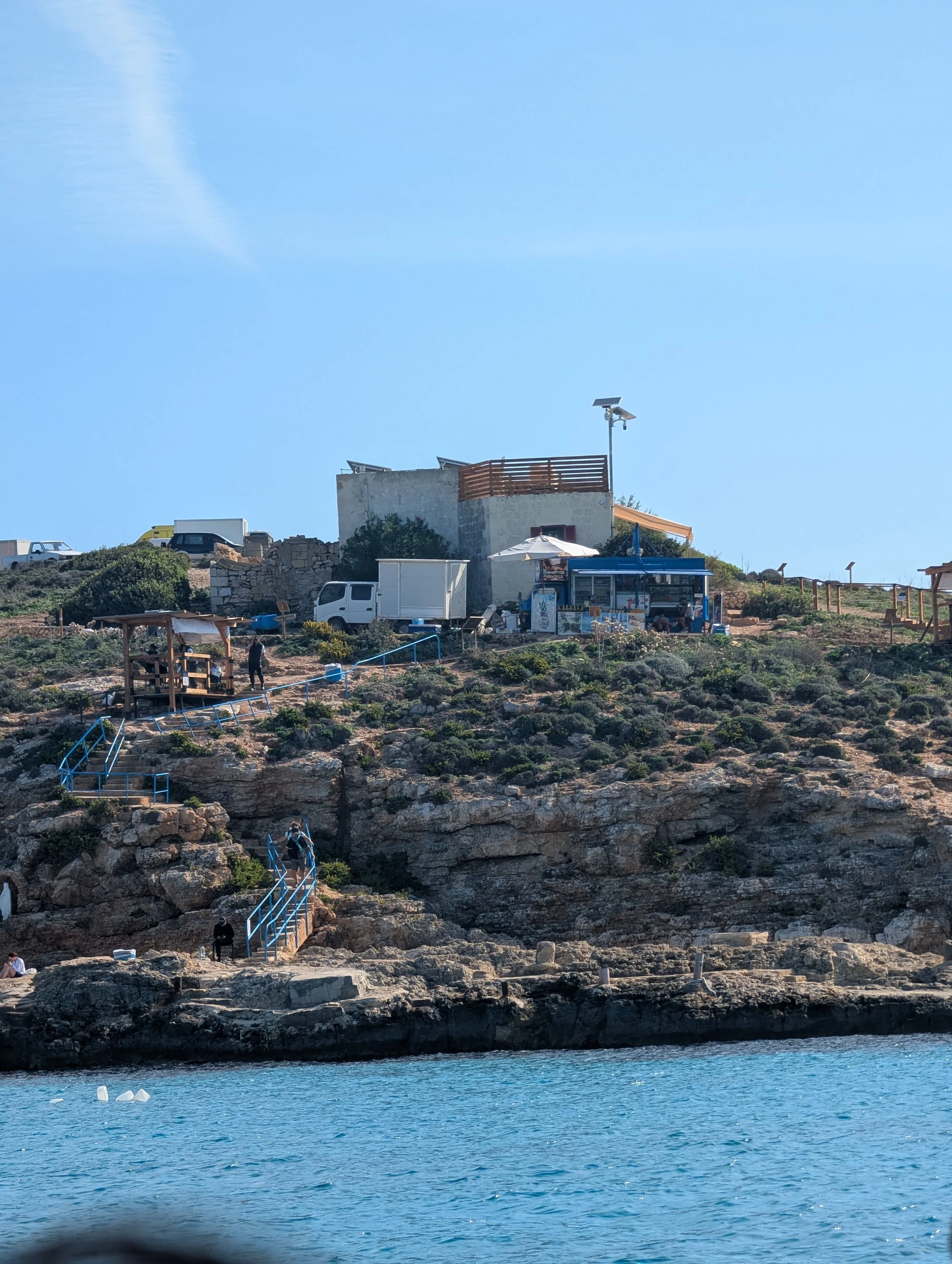 View of a rocky coastline with a blue water body in the foreground and a small building and vehicles on a hilltop in the background, under a clear blue sky.