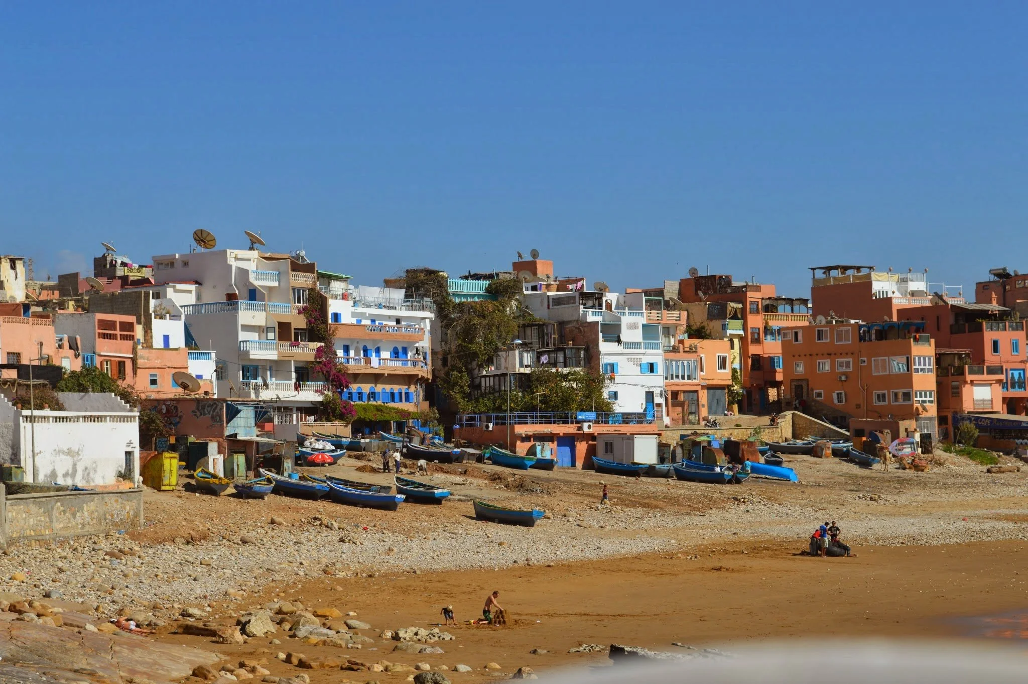 Colorful houses on a hillside near a rocky beach with boats and people, under a clear blue sky.