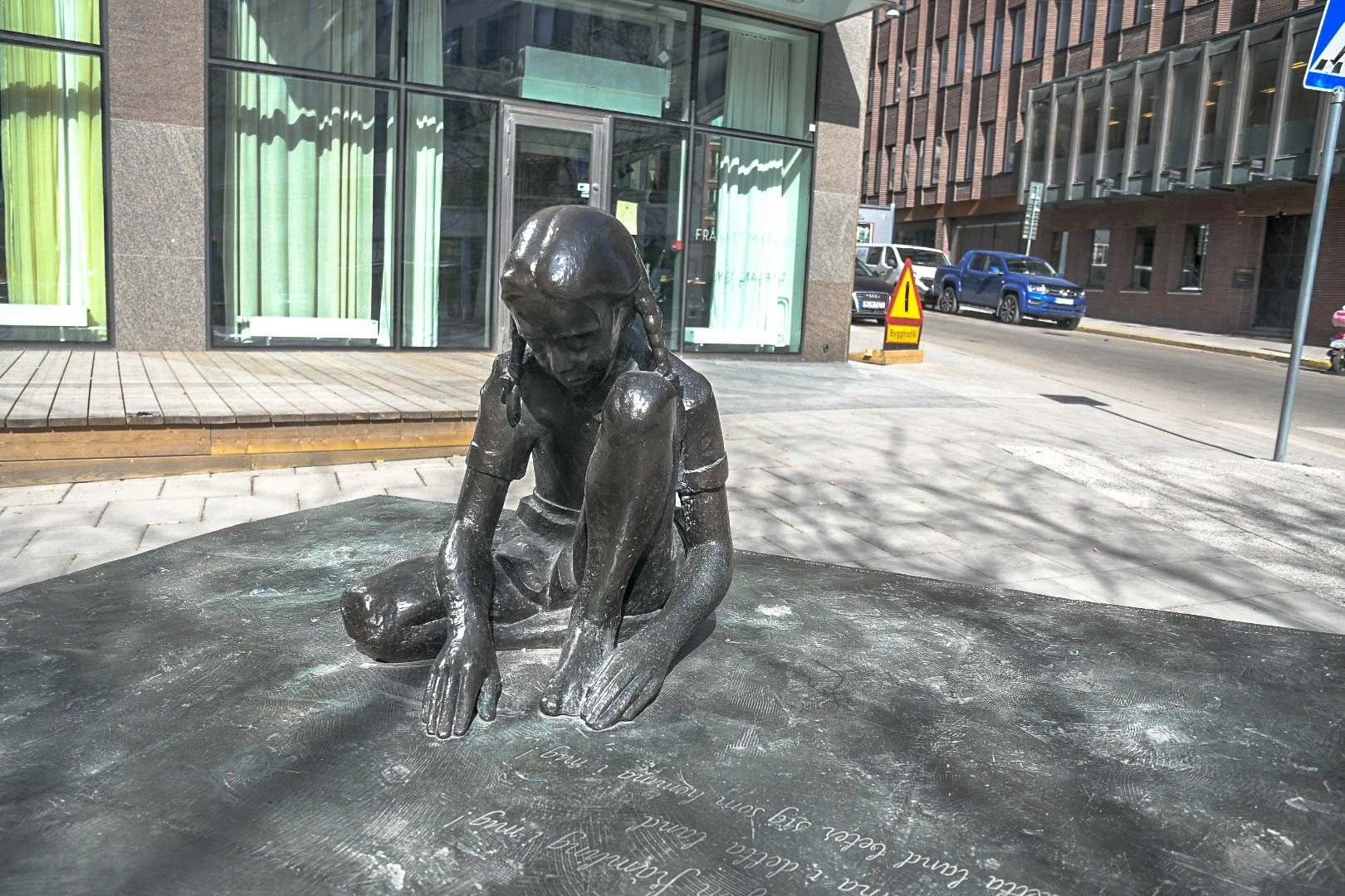 Bronze sculpture of a young girl sitting on the ground, resting her head on her knee in front of a building with glass windows and green curtains in an urban setting.
