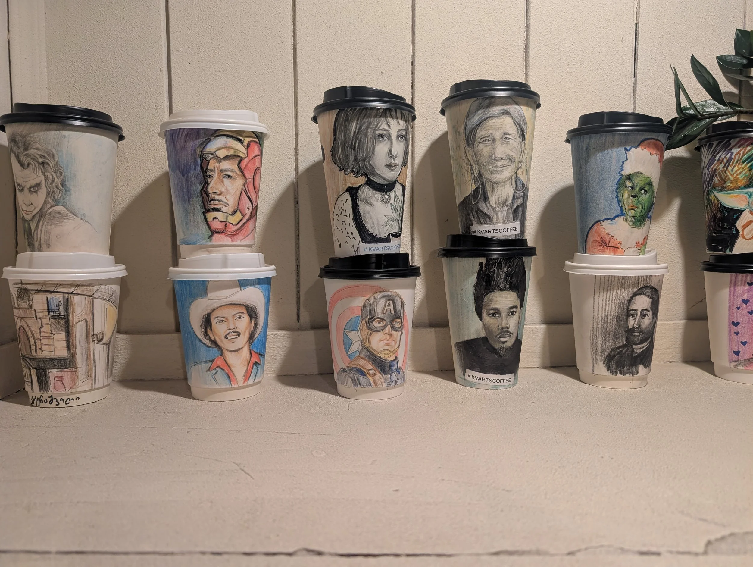 Row of disposable coffee cups with hand-drawn portraits of various famous characters and people, placed against a beige wall.