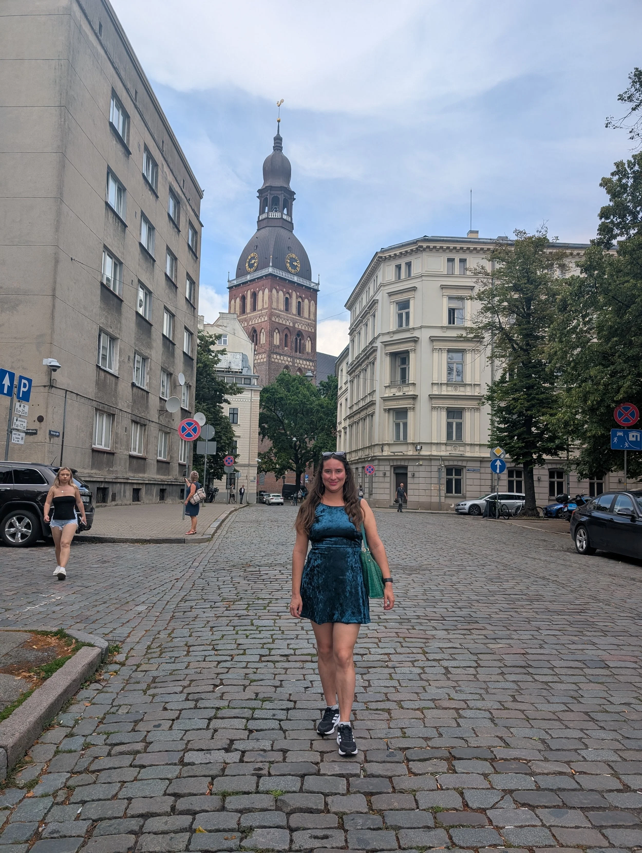 A woman in a teal velvet dress and black sneakers walking on a cobblestone street, with others in casual clothes behind her. The scene features historic buildings with ornate architecture, and a church steeple with a clock in the background, under a 