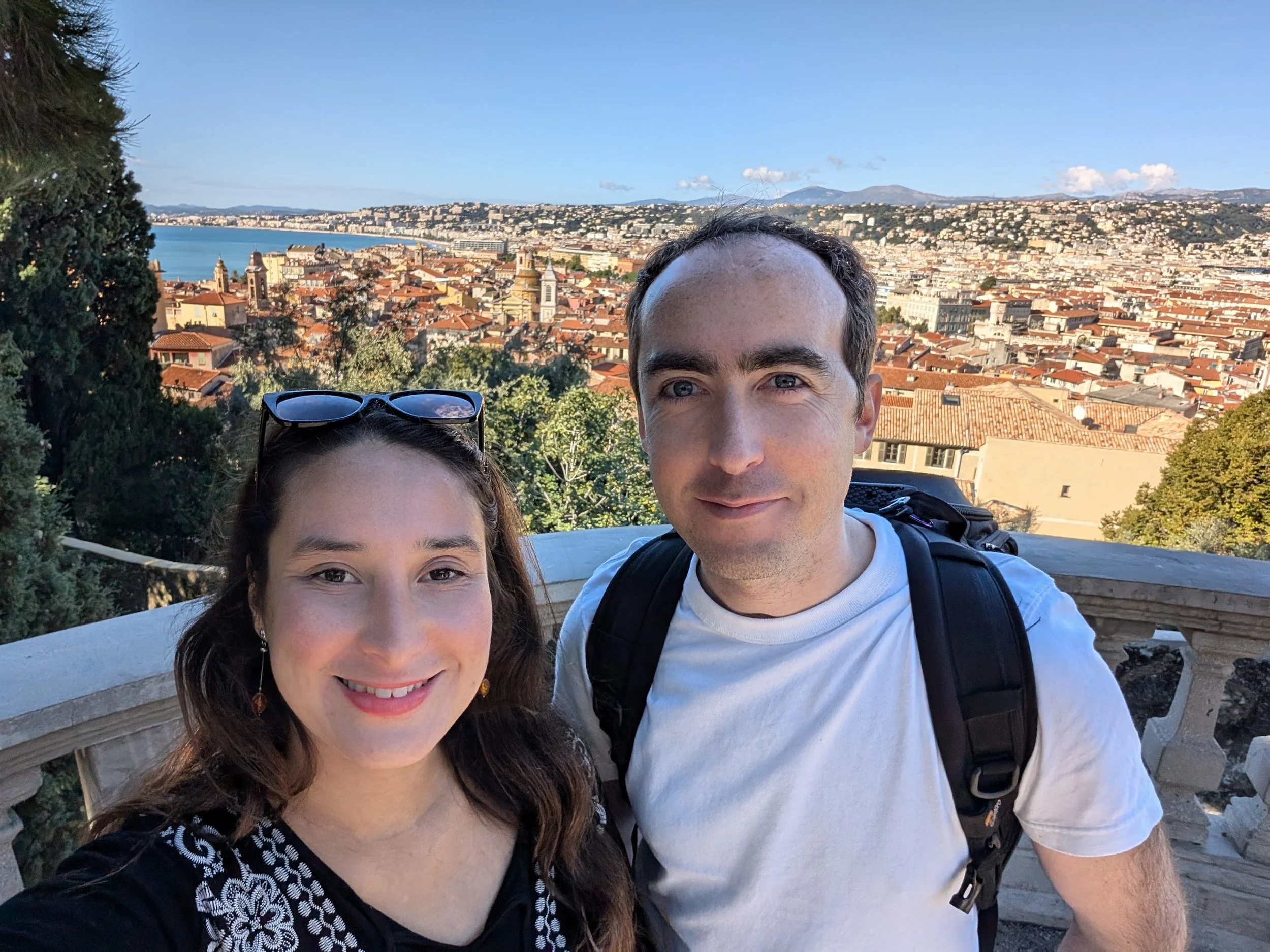 Tamara has long brown hair and is with Tom, who has short dark hair and blue eyes, taking a selfie together. They are outdoors with a cityscape, water, and mountains in the background on a clear day.