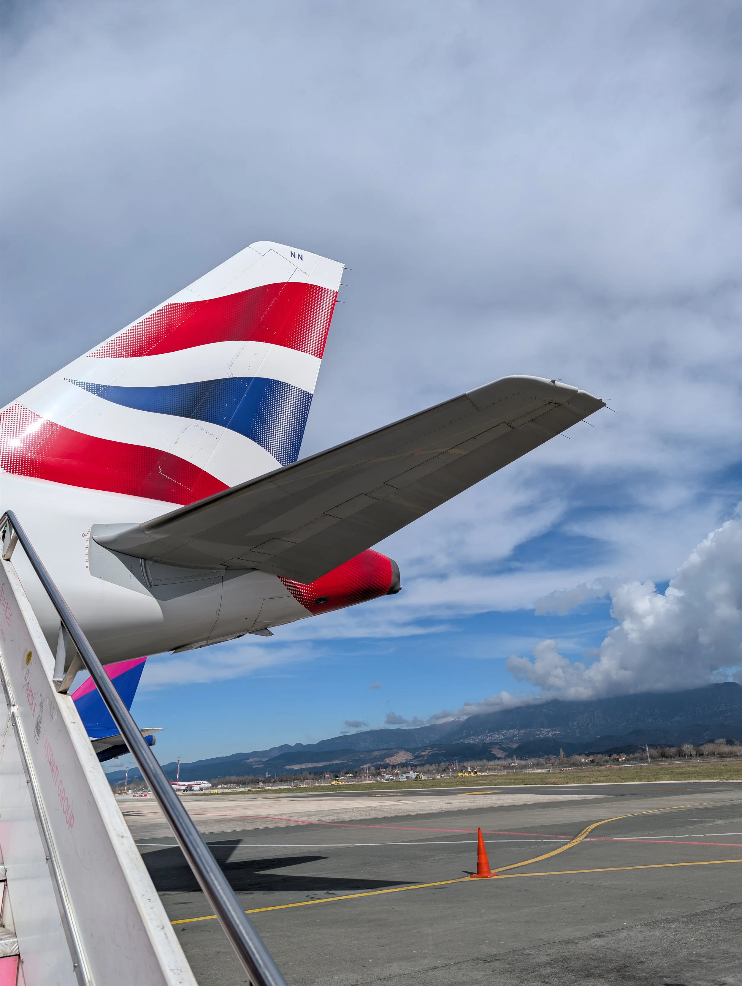 View of an airport runway with airplane tails, including a British Airways tail, and a cloudy sky in the background.