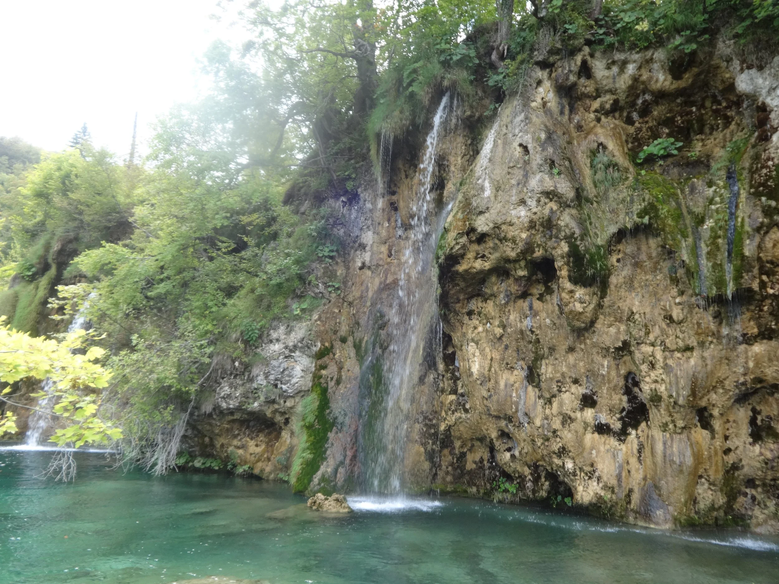 A scenic view of a waterfall flowing over rocks into a calm green pool surrounded by lush trees.