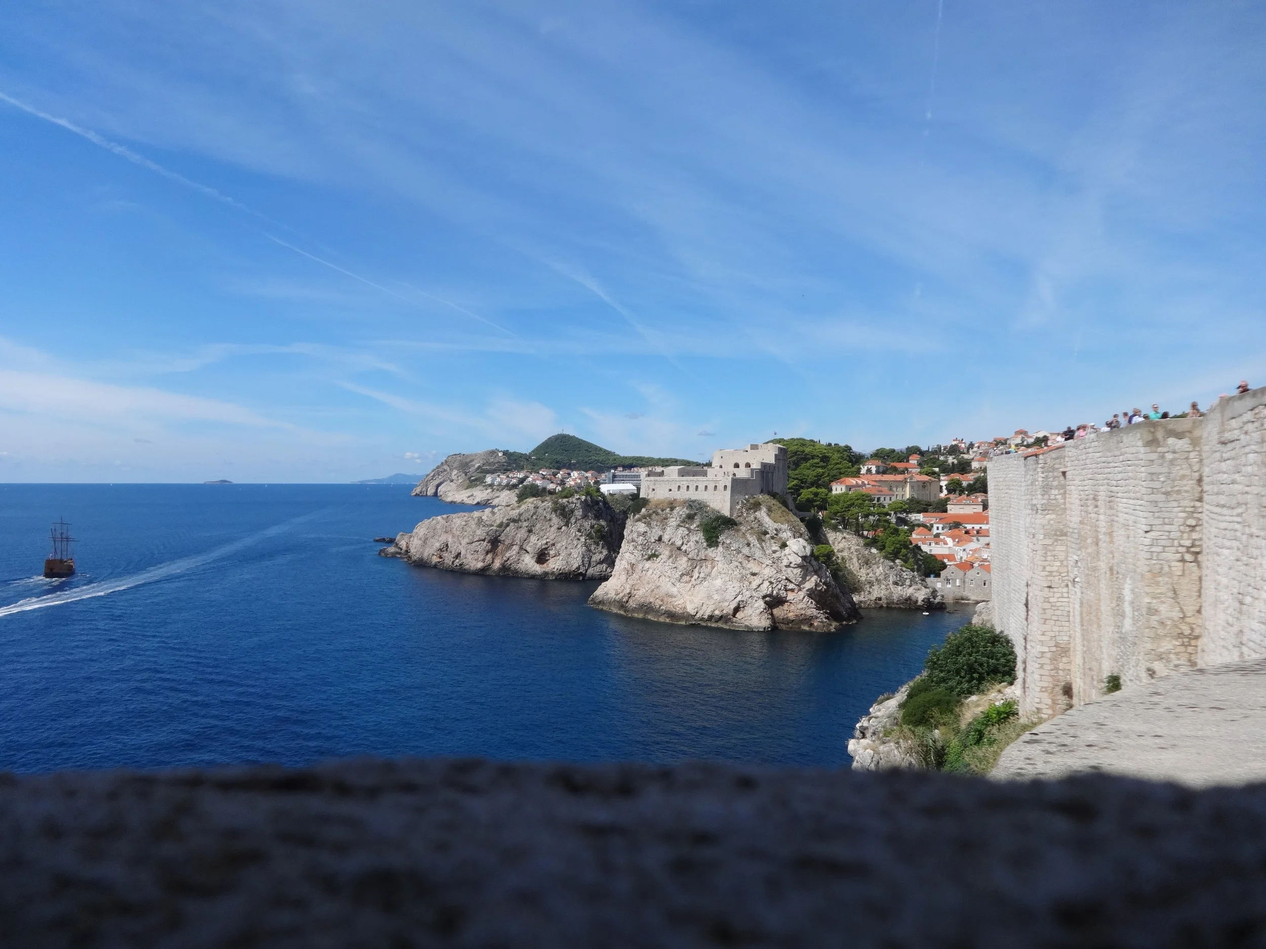 A scenic view of a coastal area with blue water, rocky cliffs, a fortress with stone walls, and houses with orange roofs on a hillside under a blue sky with wispy clouds.