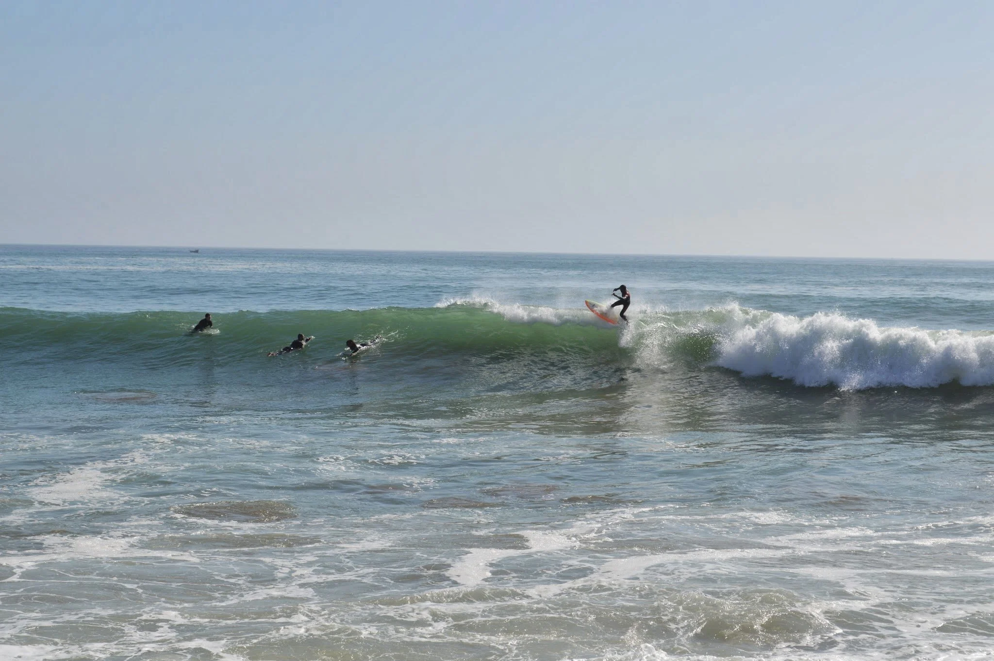 A surfer riding a wave at the beach with three other surfers in the water, and a boat visible in the distance on the horizon.