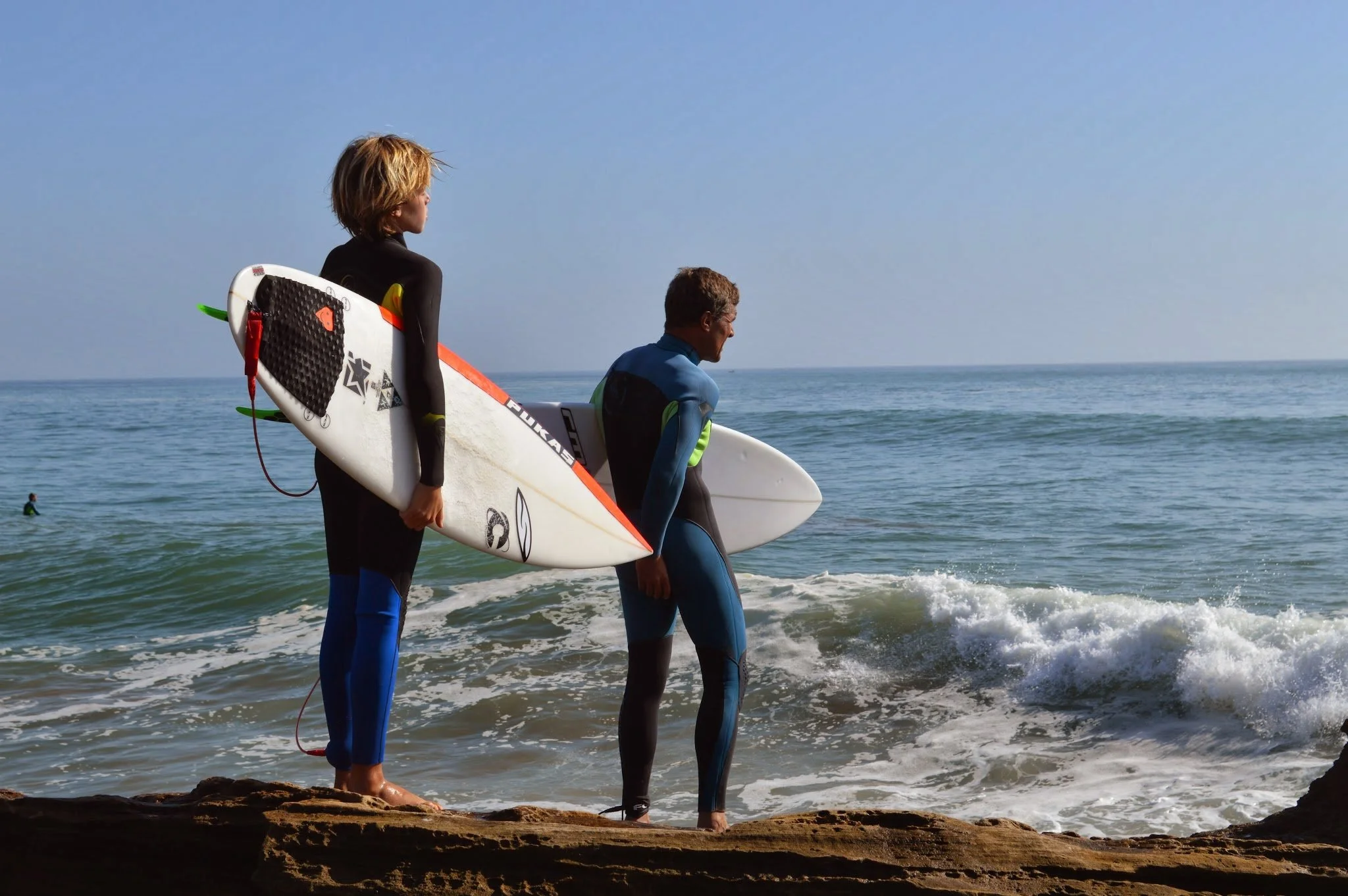 Two surfers walking along the shoreline with surfboards under their arms, looking at the ocean on a sunny day.
