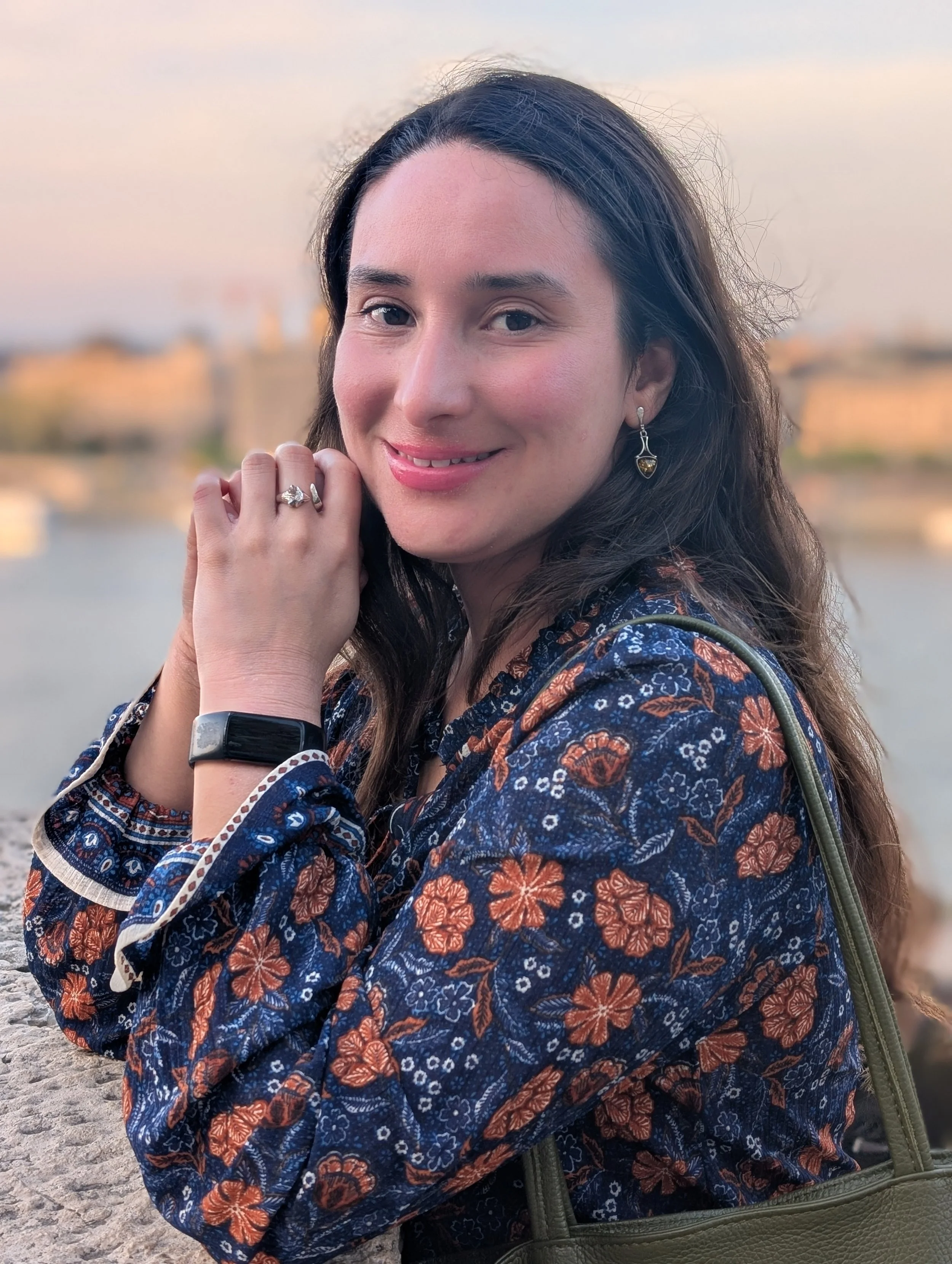 A woman with long dark hair smiling at the camera, wearing earrings, a patterned dress, a ring, and a smartwatch, with a blurry outdoor background.