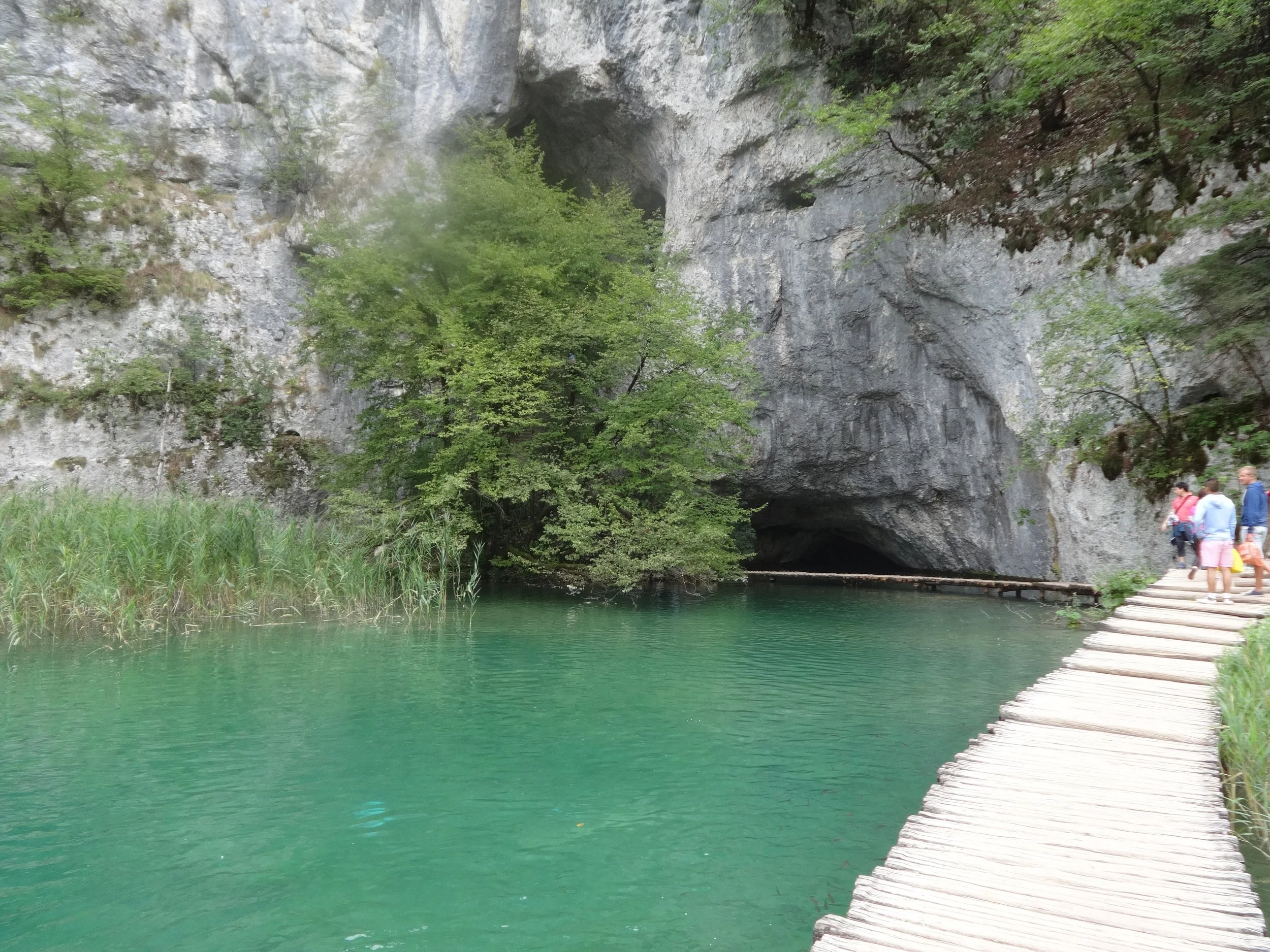 People walking on a wooden pathway alongside a green lake with a cliff and a cave entrance in the background.