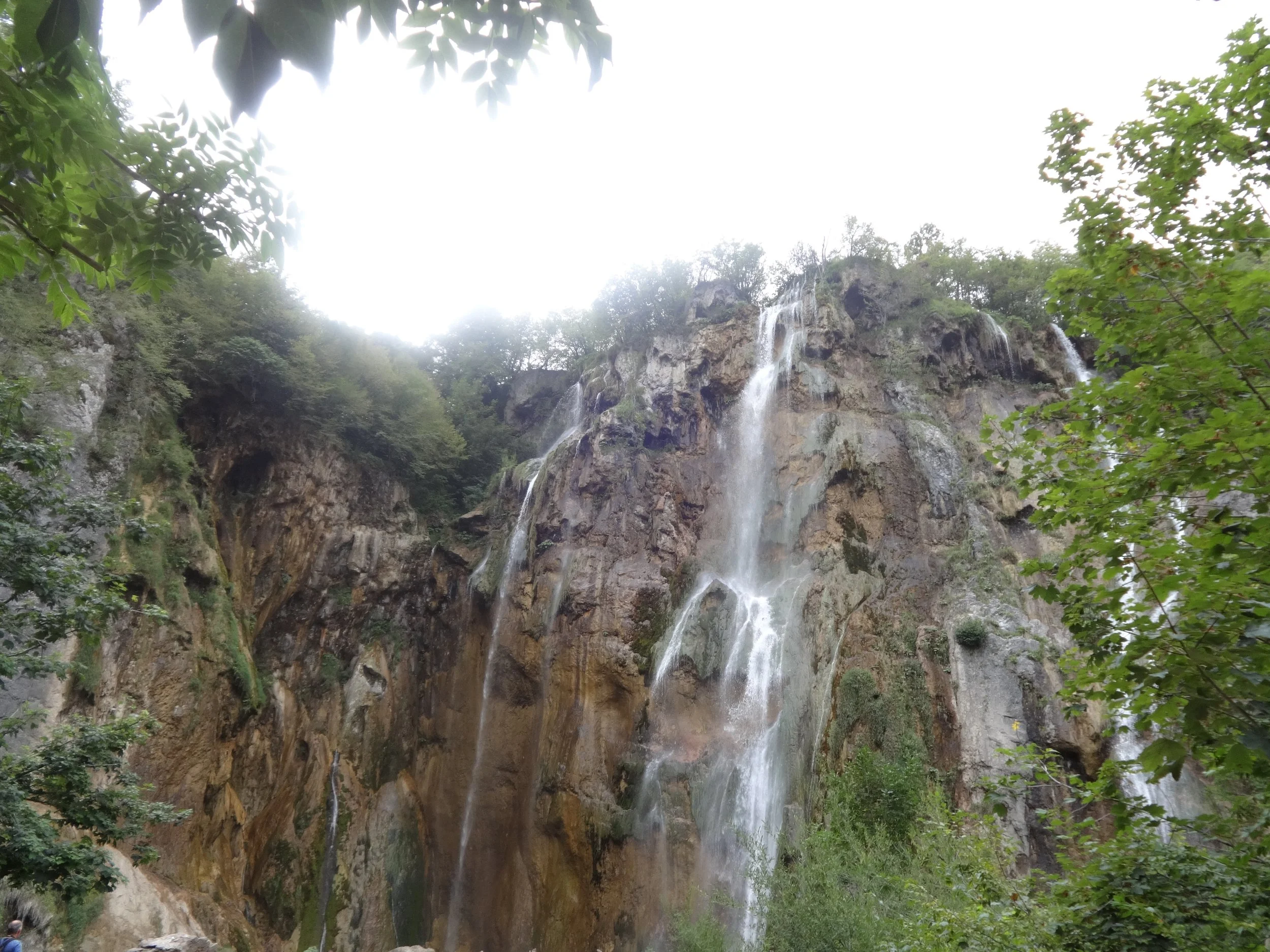 Tall waterfall cascading down a rocky cliff surrounded by green trees and foliage.