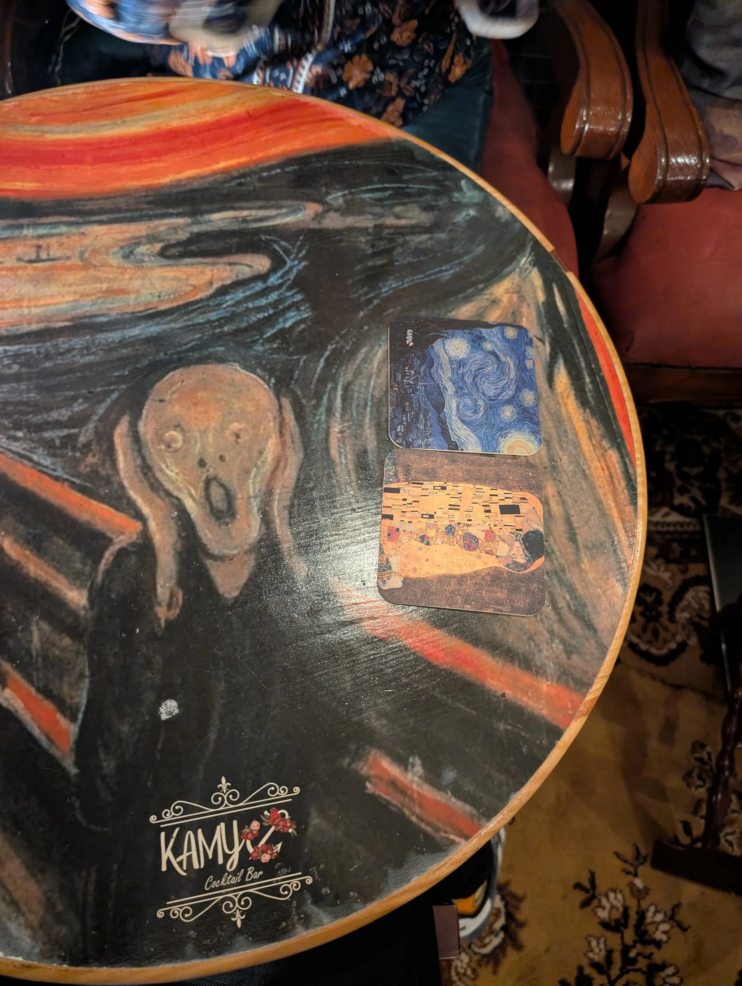 A round table with a black top featuring a vibrant swirling pattern of red, orange, and black. On the table, there are two cards with artwork inspired by Vincent van Gogh's "Starry Night" and Gustav Klimt's "The Kiss." In the background, a person in 