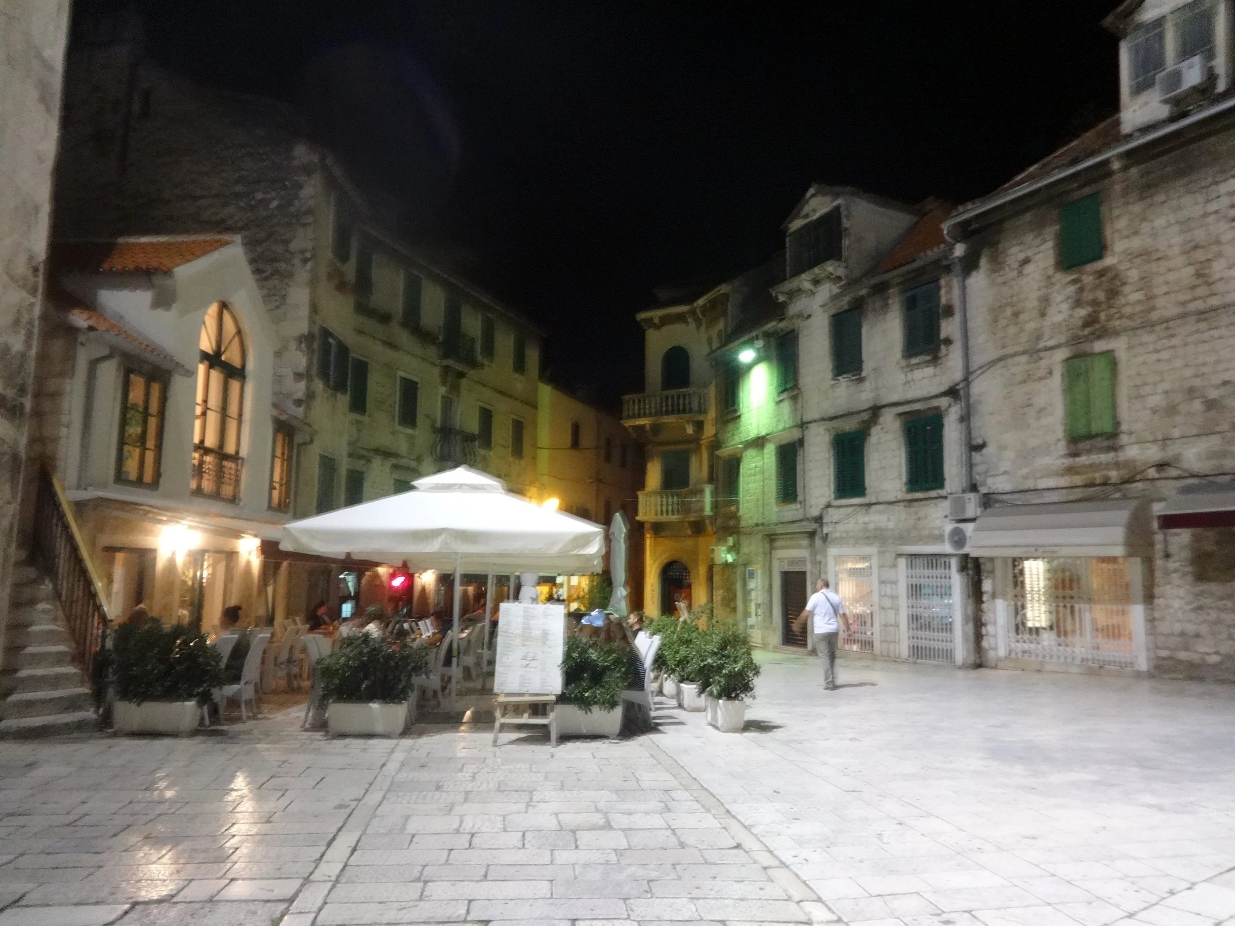 Nighttime scene of a small European town square with outdoor seating, tables with umbrellas, and historic stone buildings with green shutters, some with balconies, illuminated by street lights.