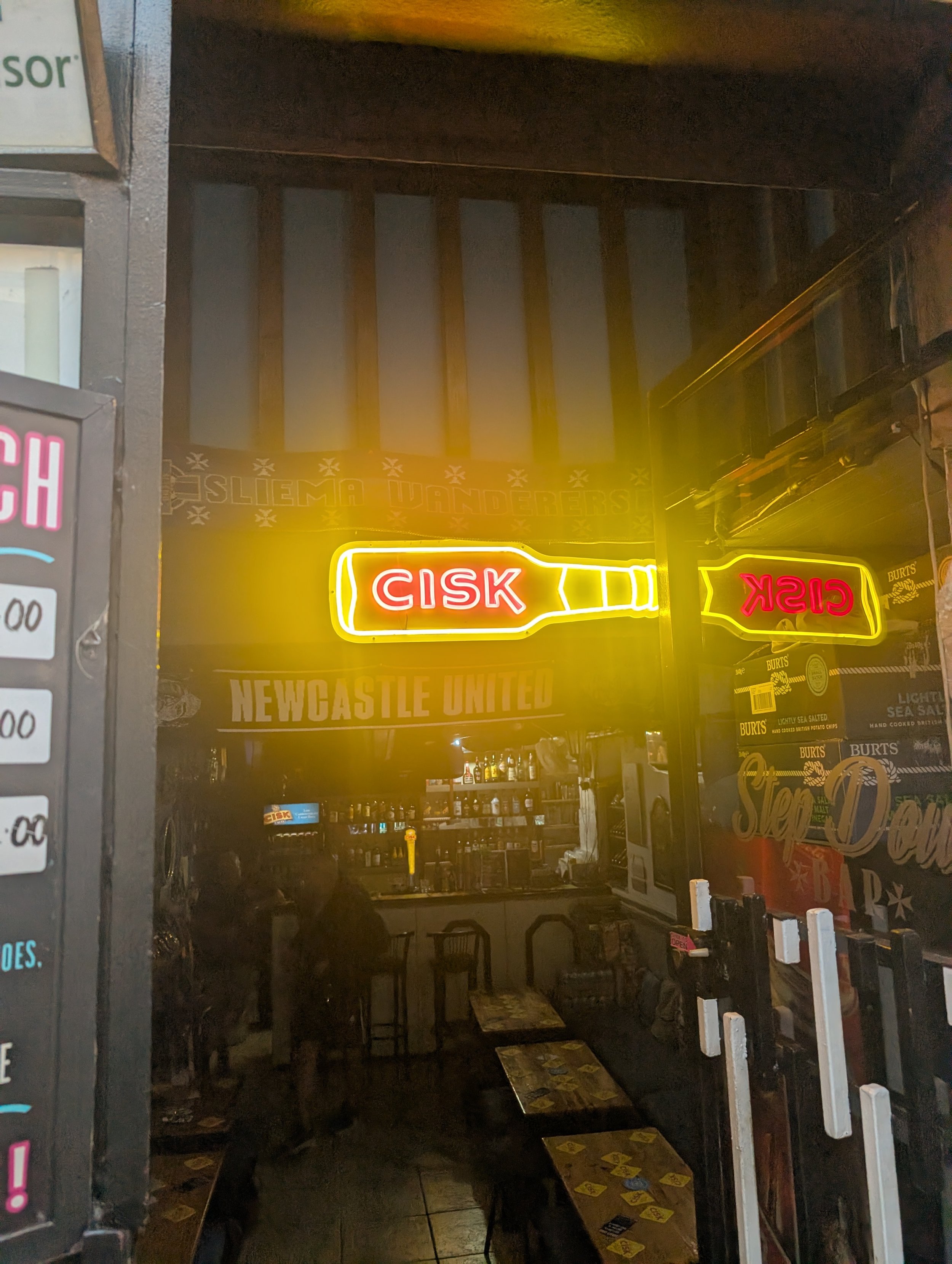 Interior of a dimly lit restaurant or bar, with a neon sign reading 'CISK' in yellow and red, tables and chairs, and a bar counter visible in the background.