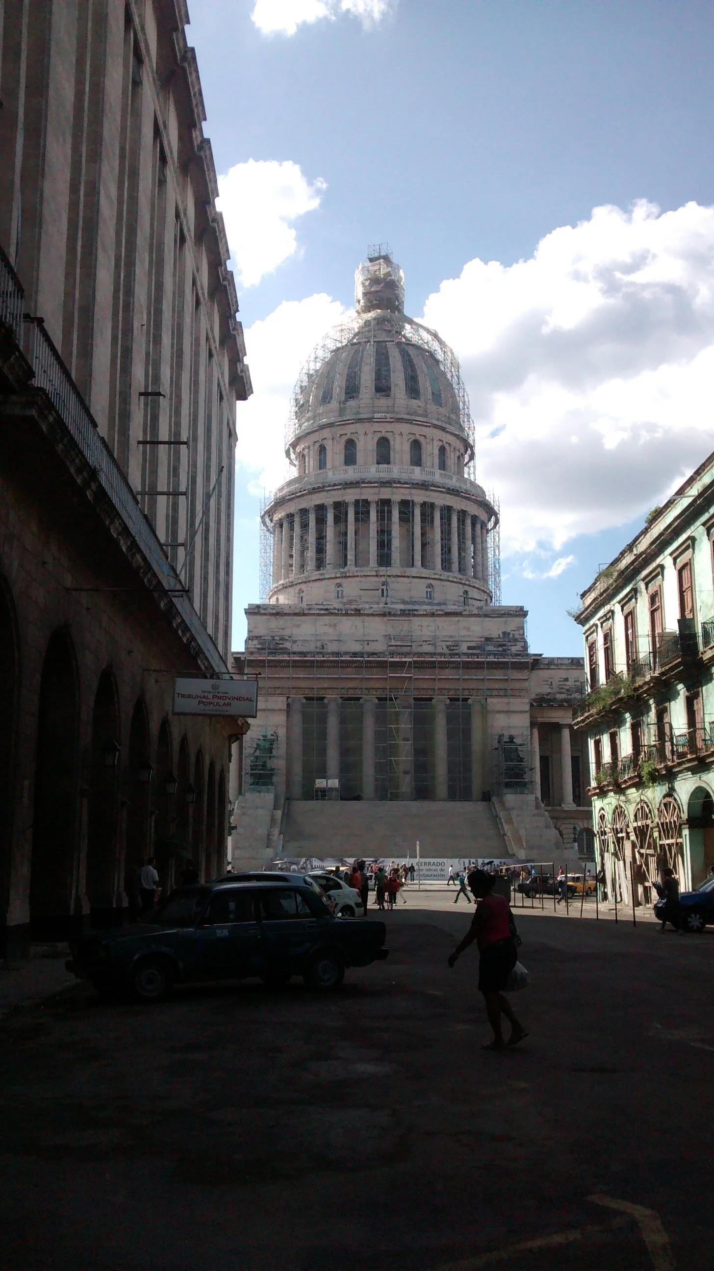 View of a historic building with a dome under renovation, scaffolding surrounding the upper part, located at the end of a street with cars parked and people walking, under a partly cloudy sky.