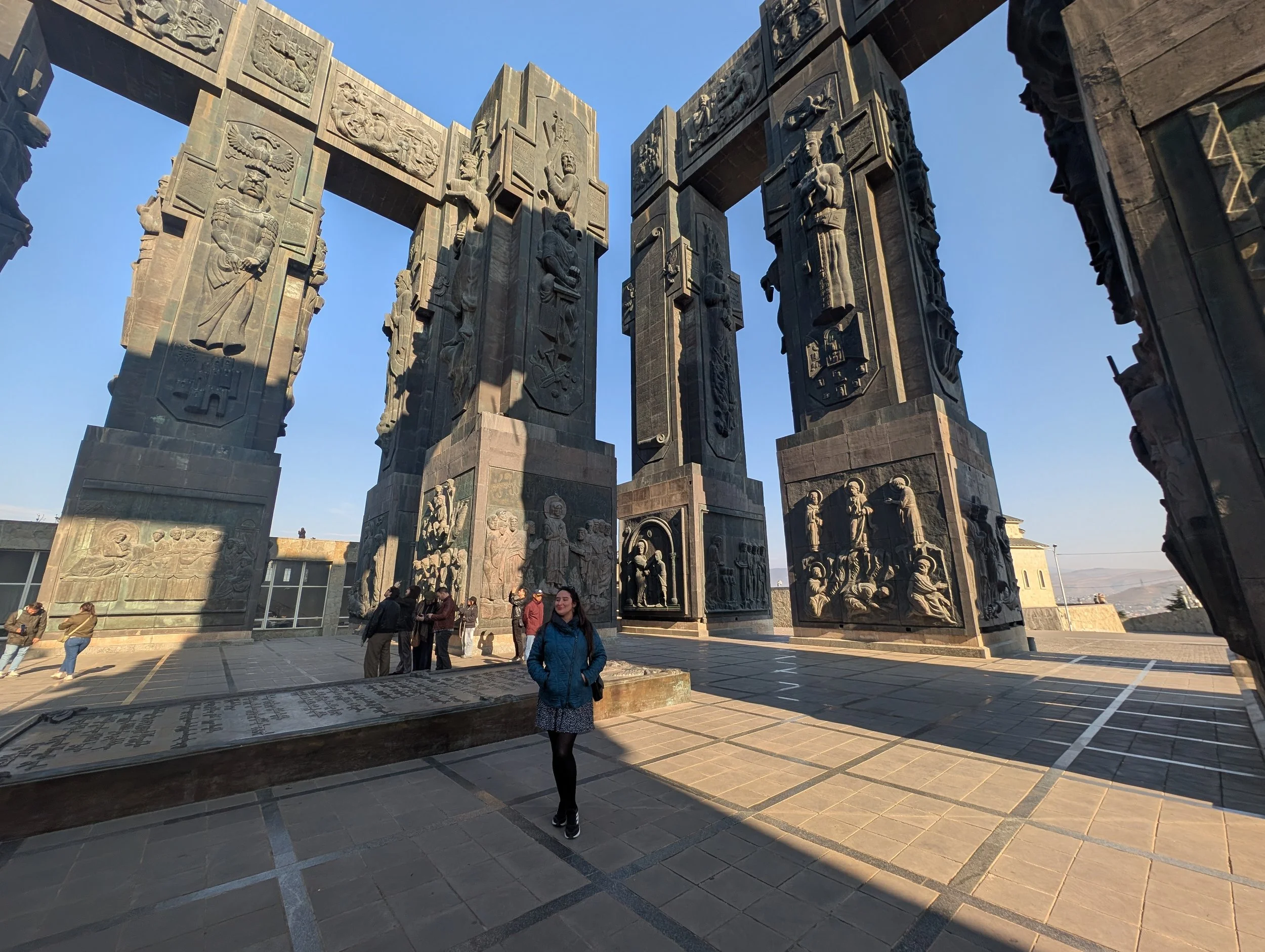 View of the Black Wall Street Memorial with large stone pillars engraved with various historical figures and symbols, and several people walking and standing nearby on a paved area under a clear blue sky.