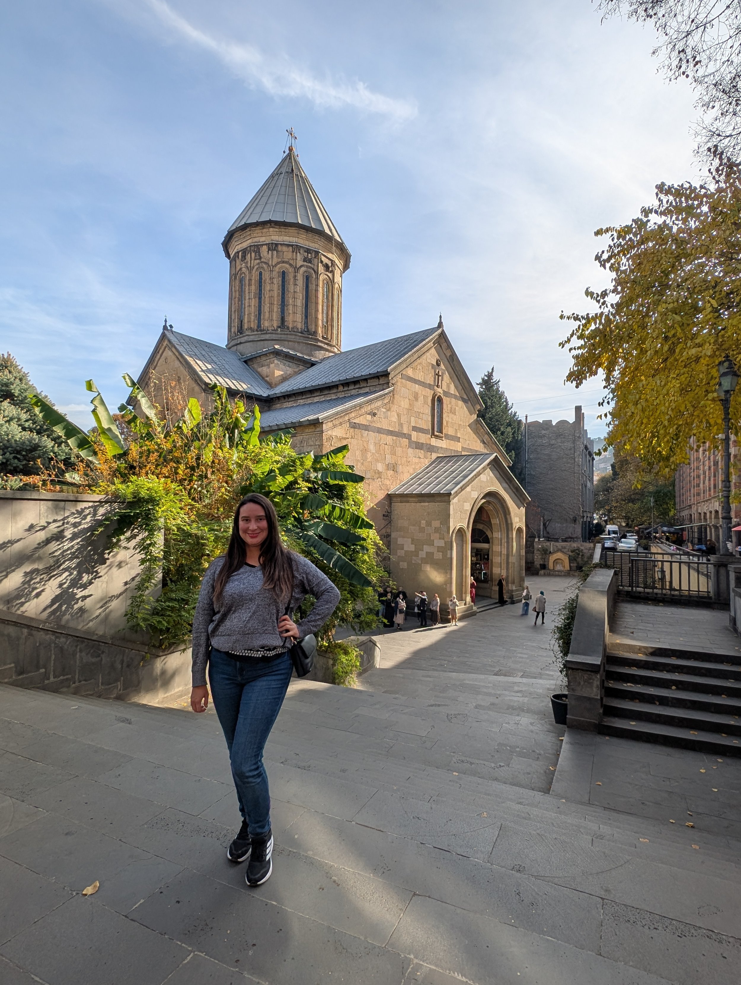 A woman with long dark hair, wearing a grey sweater, blue jeans, and black sneakers, stands on a stone-paved street in front of a historic stone church with a tall, pointed steeple. There are trees with green and yellow leaves, people walking in the 