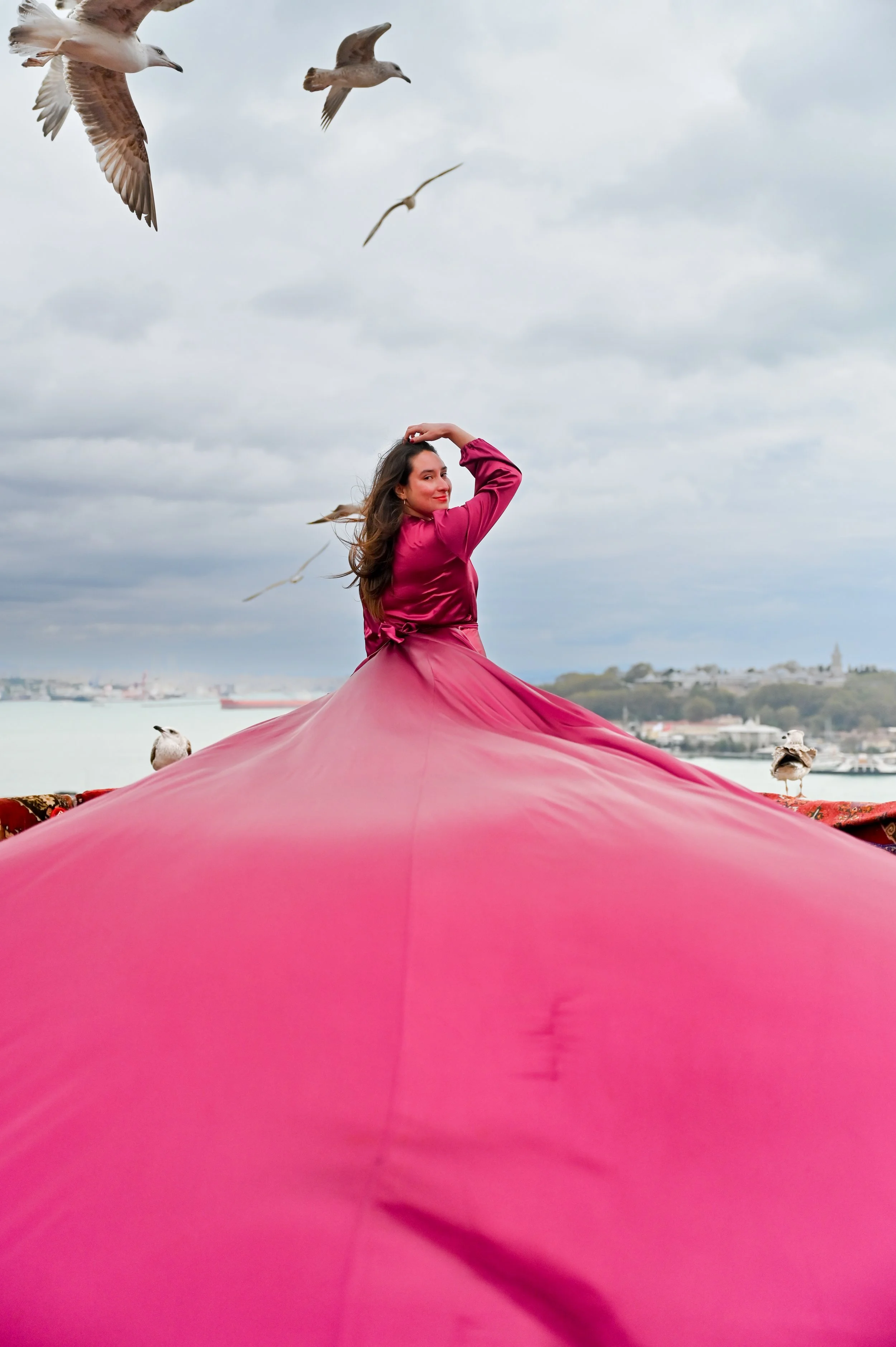 A woman in a pink dress standing on a colorful platform near water, with seagulls flying in the cloudy sky behind her.