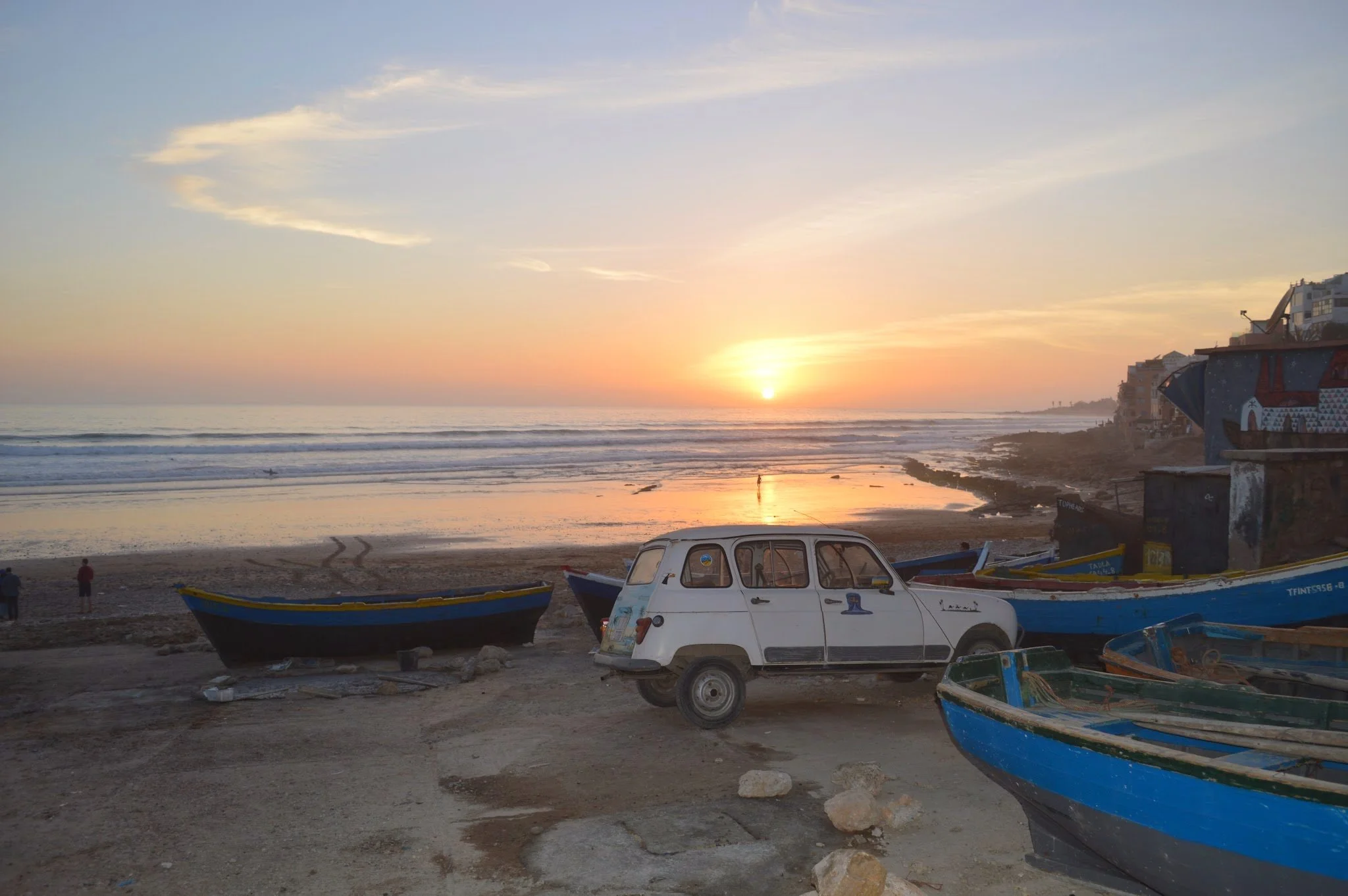 Sunset over the ocean with boats on the beach, buildings on the right, and a car in the foreground.