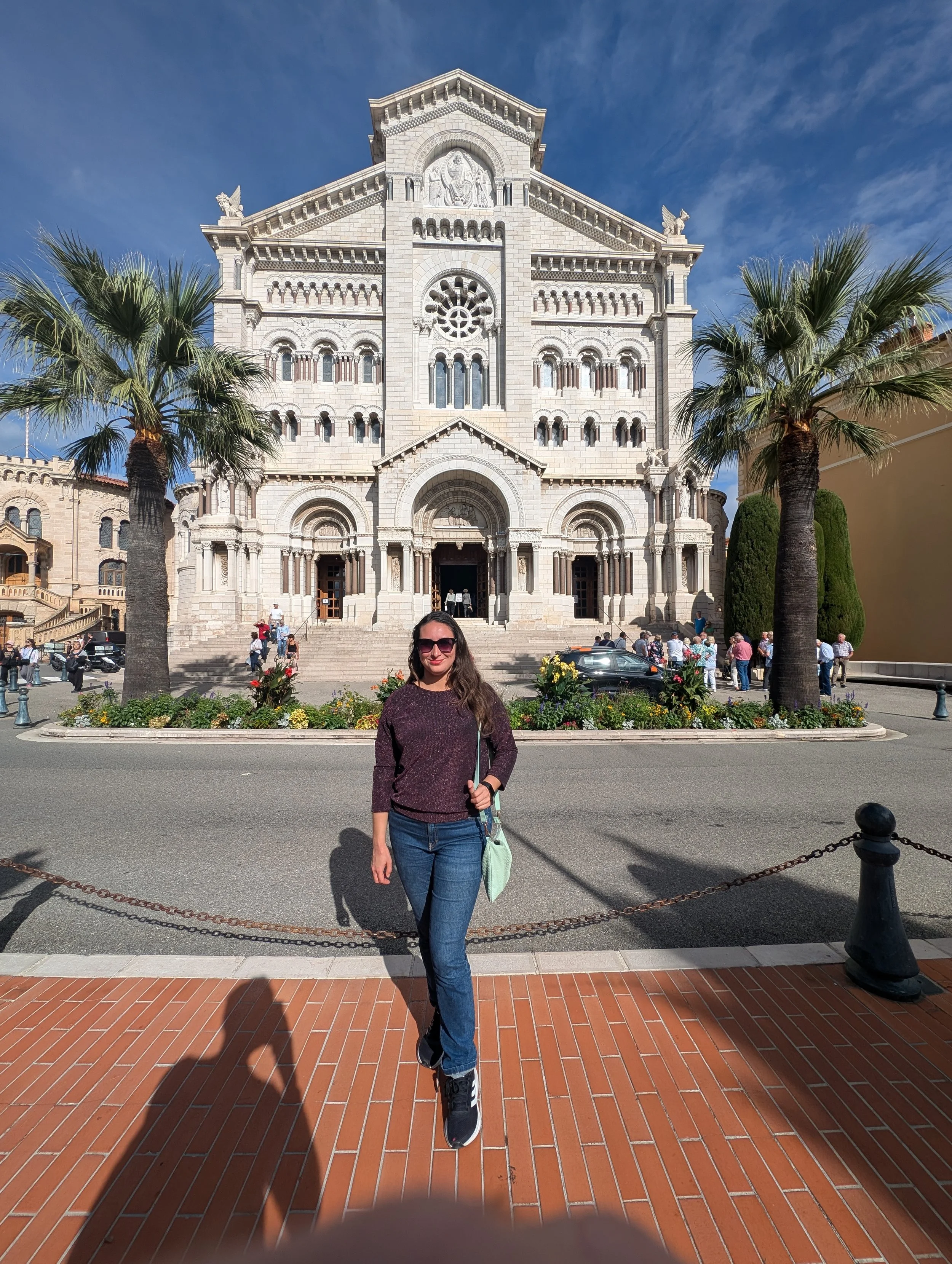 A woman with long brown hair, sunglasses, a purple sweater, and blue jeans stands on a brick sidewalk in front of a large cathedral with a stone facade, steps, and palm trees.