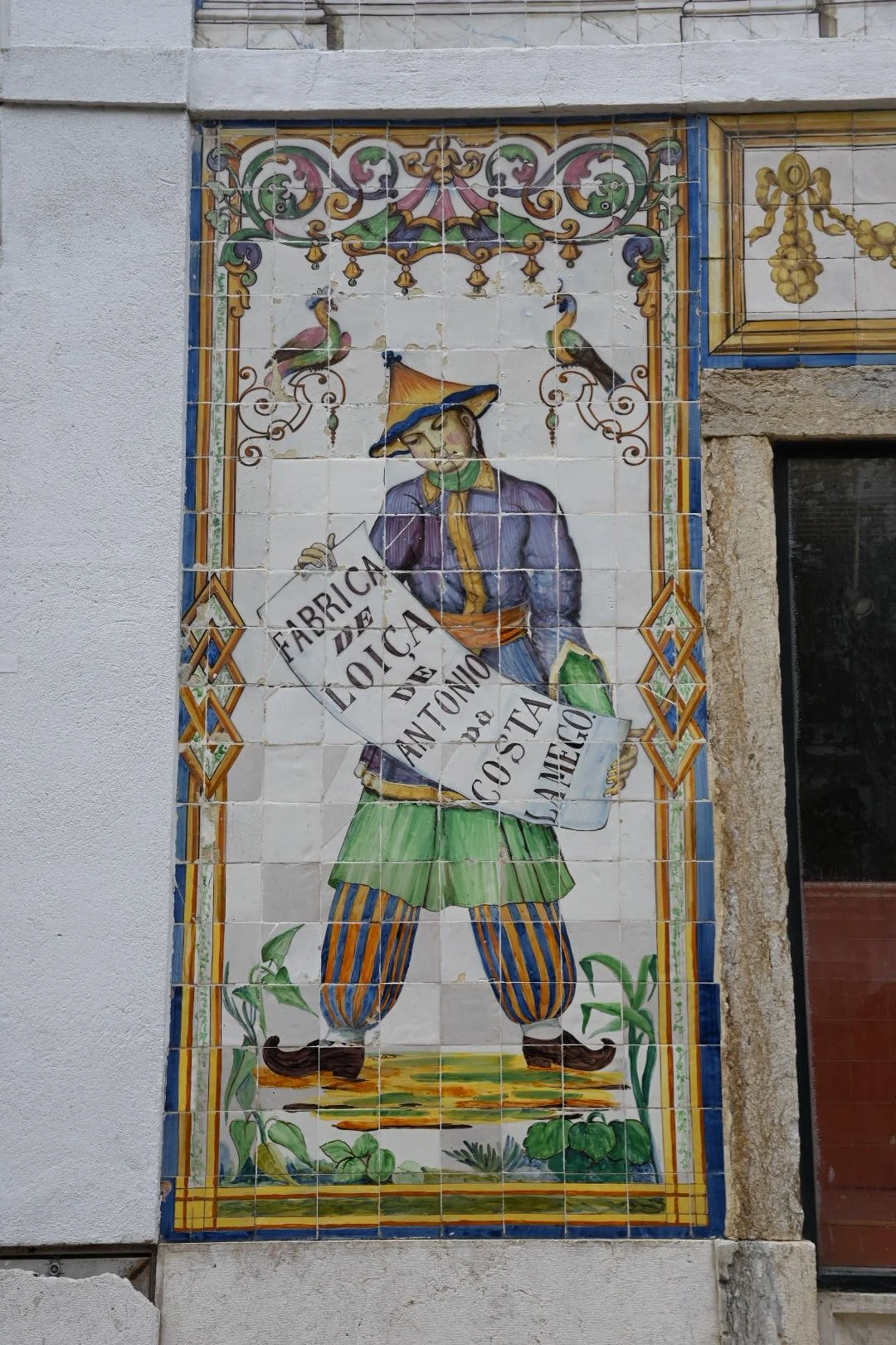 Colorful traditional Portuguese tile mural depicting a man in period costume holding a sign that reads 'Fábrica de Roca de António da Costa', with decorative elements like trees, birds, and floral patterns.